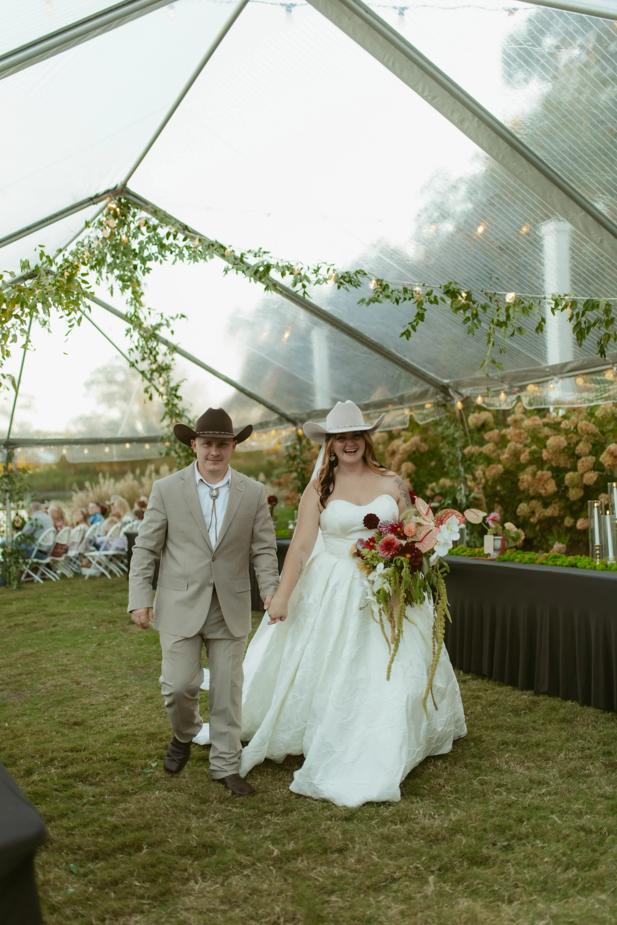 A bride and groom walking under a clear tent decorated with greenery and string lights at their wedding reception. The bride is wearing a strapless white gown and a white cowboy hat, holding a vibrant bouquet. The groom is dressed in a beige suit and a brown cowboy hat, holding the bride's hand.
