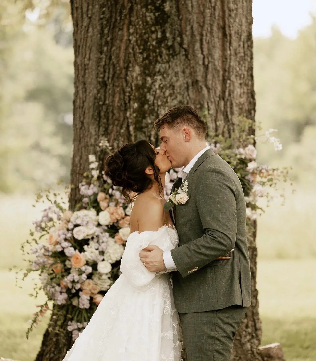 A bride and groom sharing a kiss outdoors during their wedding, with a large tree and a floral arrangement in the background.