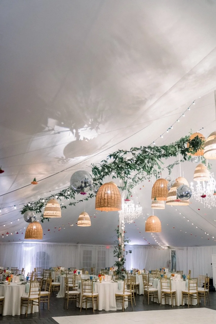Elegant event tent decorated with hanging basket lamps, greenery, string lights, and reflective disco balls, with round tables and gold chairs for a celebration.