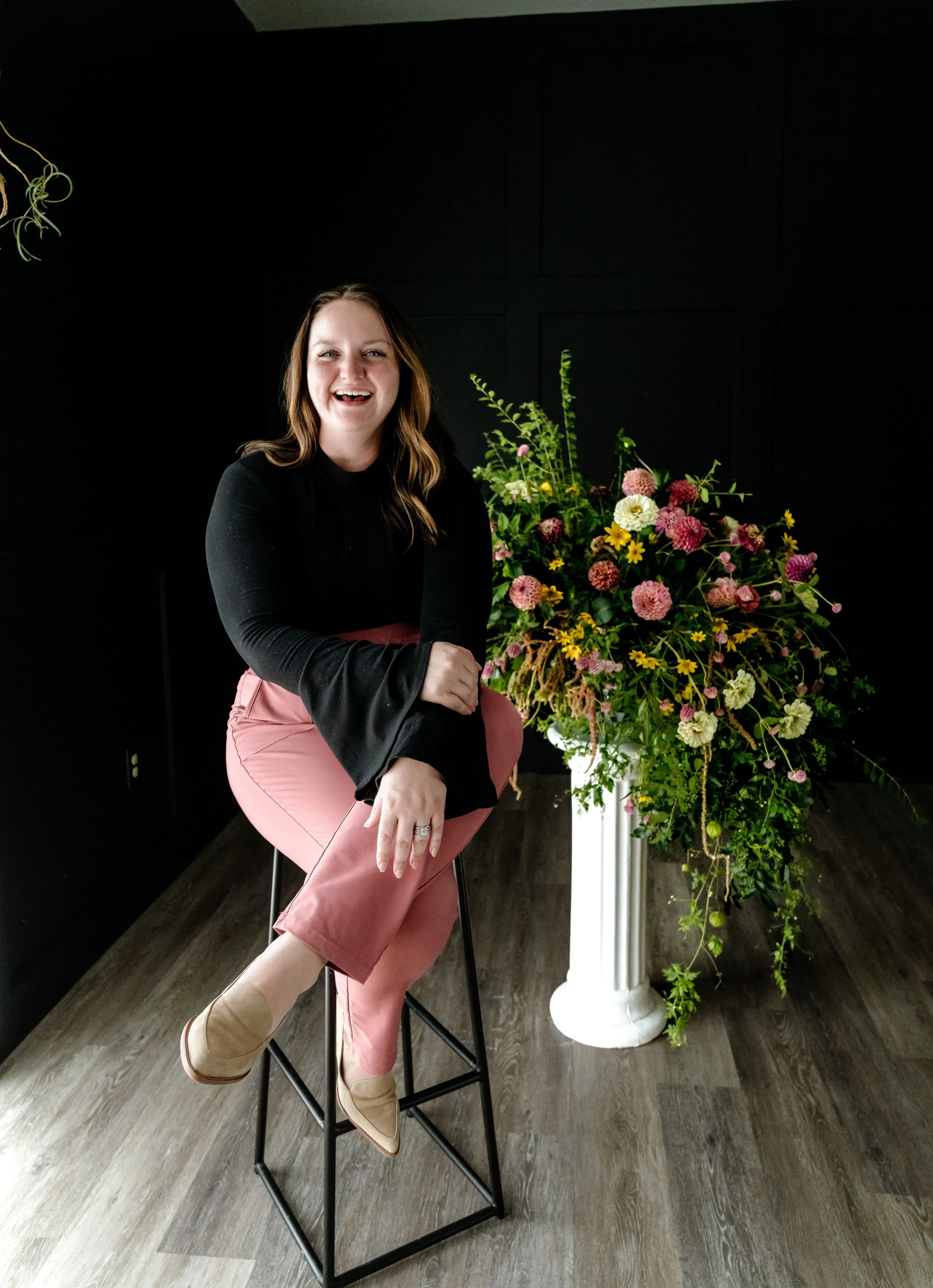 A woman sits on a black stool next to a large bouquet of colorful flowers in a white vase, in a room with dark walls and wood flooring.