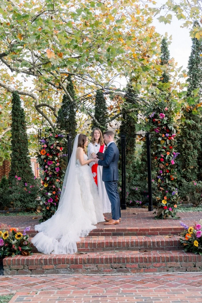 A bride and groom exchange vows at a wedding ceremony outdoors under a black arched structure decorated with colorful flowers, with a female officiant standing behind them, in front of trees with green and yellow leaves.
