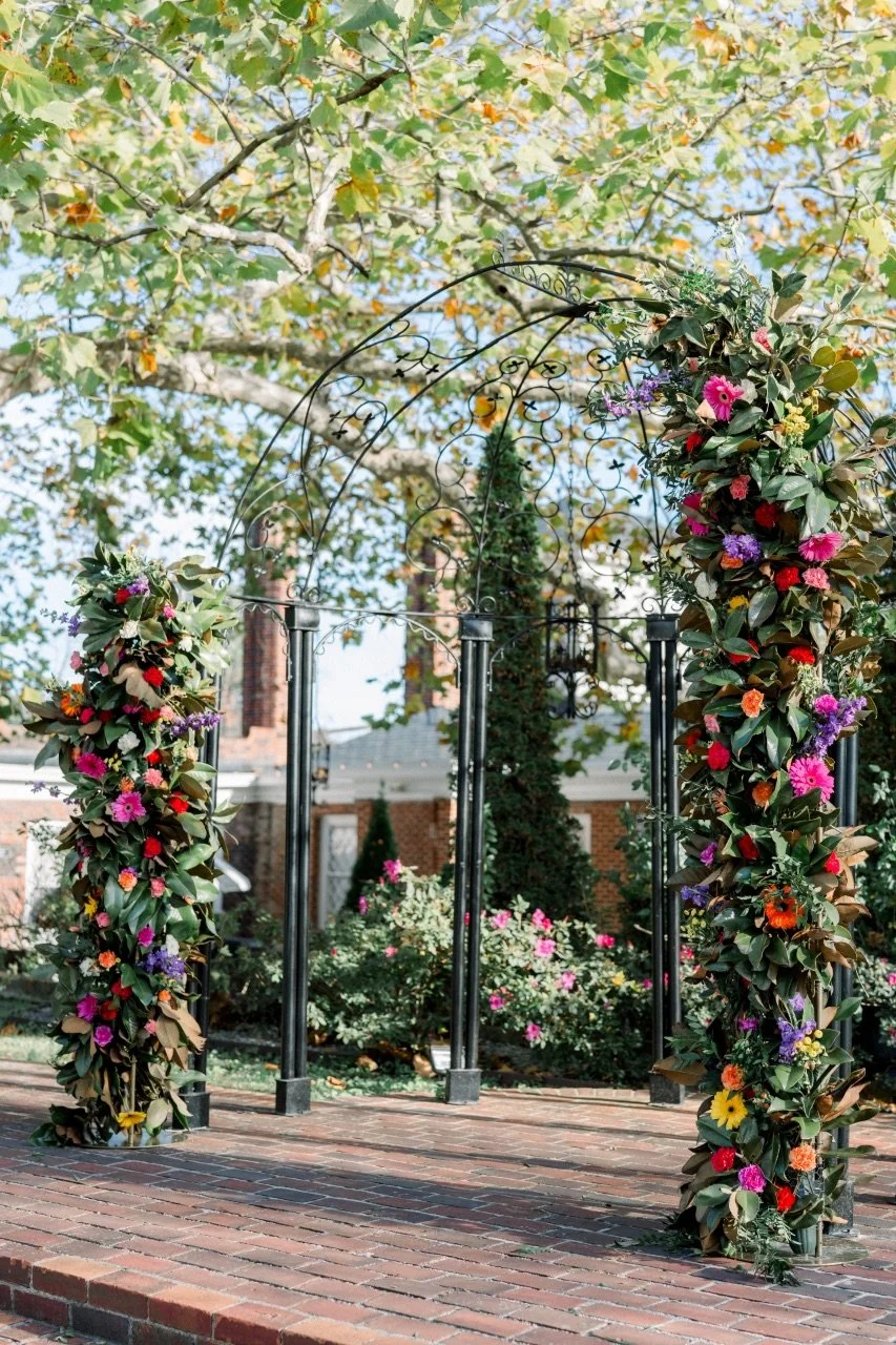 An ornate iron garden gate decorated with colorful flowers, surrounded by leafy trees and flowering bushes in a garden setting.