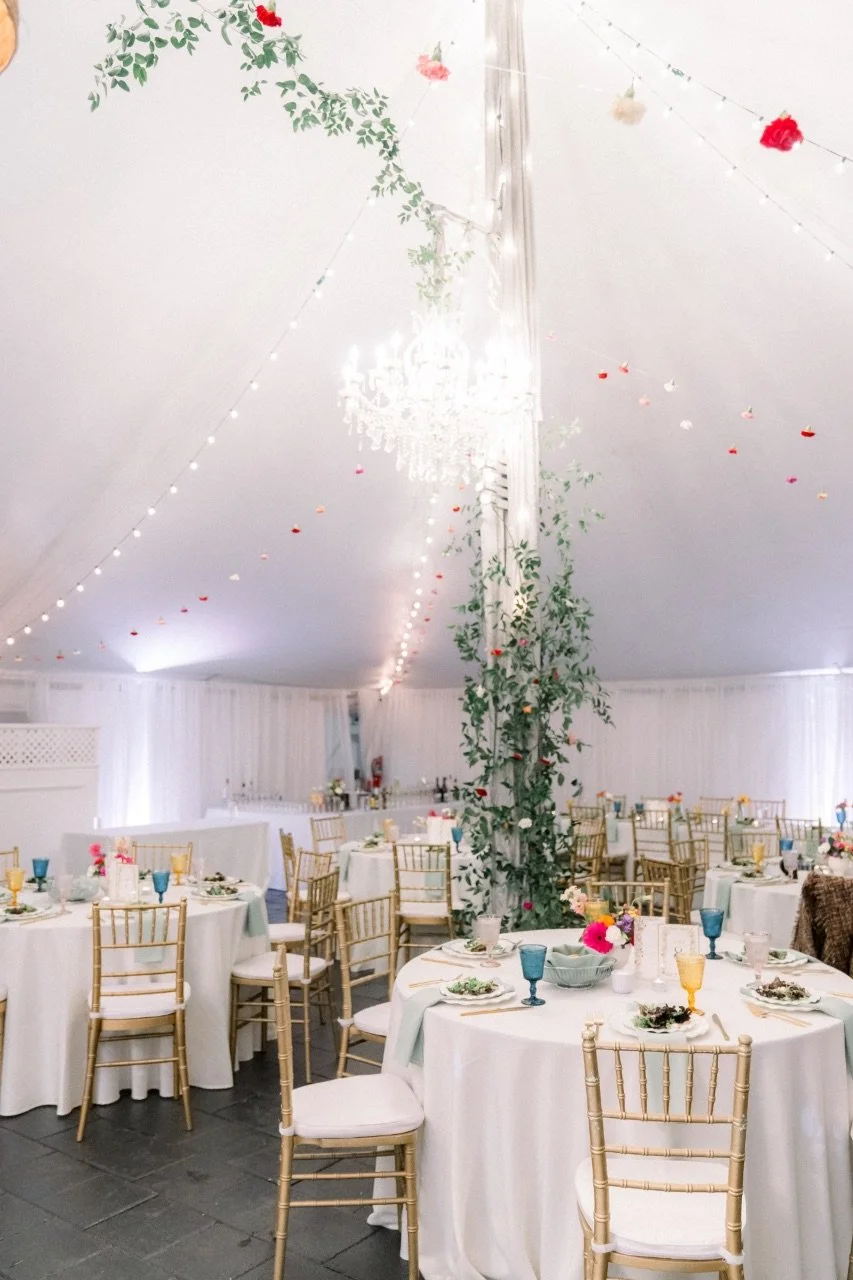 Elegant event tent with white draped walls, decorated with string lights and hanging floral garlands. Round tables with white tablecloths, gold chairs, and colorful glassware are set for a celebration.