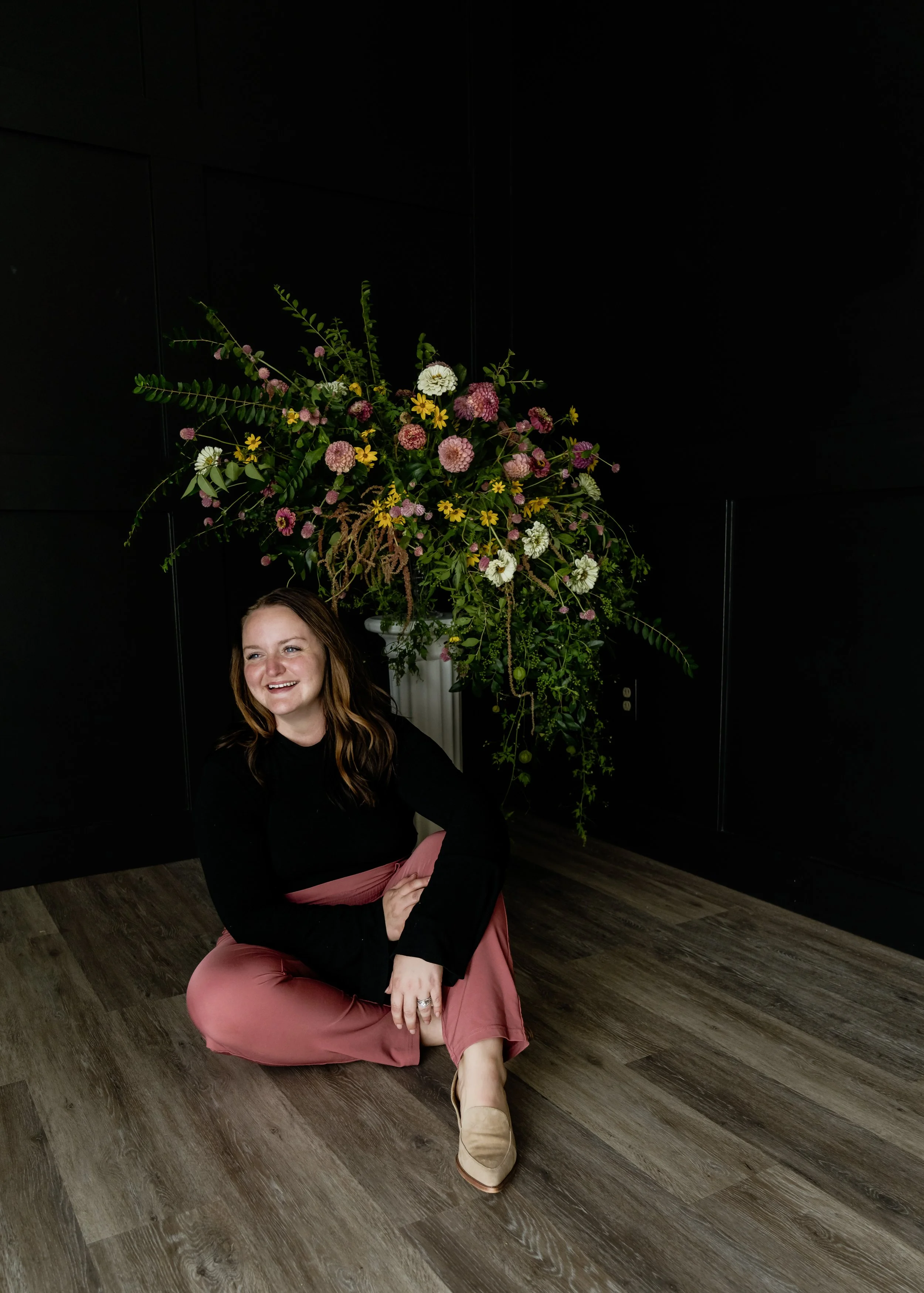 A woman sitting on the floor smiling, with a large floral arrangement in a white vase behind her, set against a black wall.