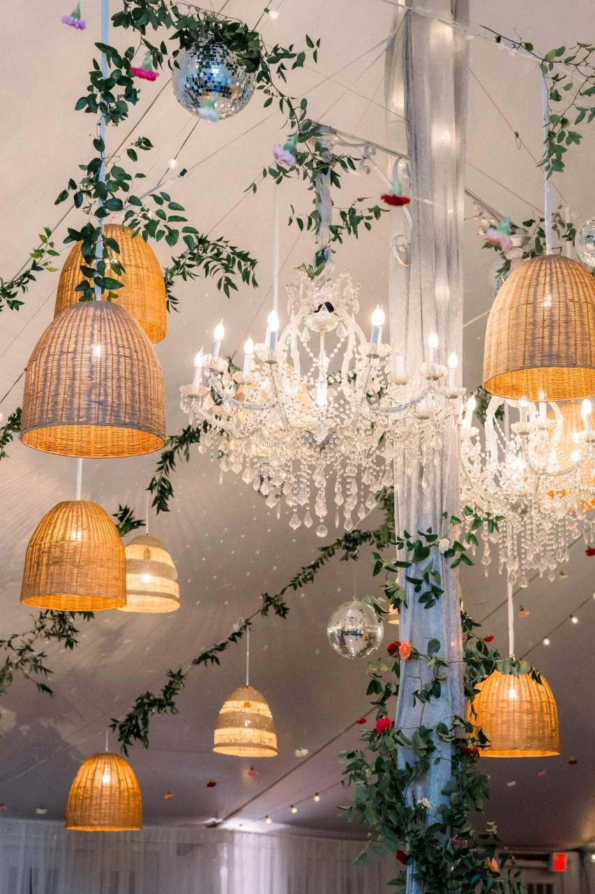Decorative ceiling arrangement featuring chandeliers, hanging woven lamps, string lights, greenery, and disco balls.