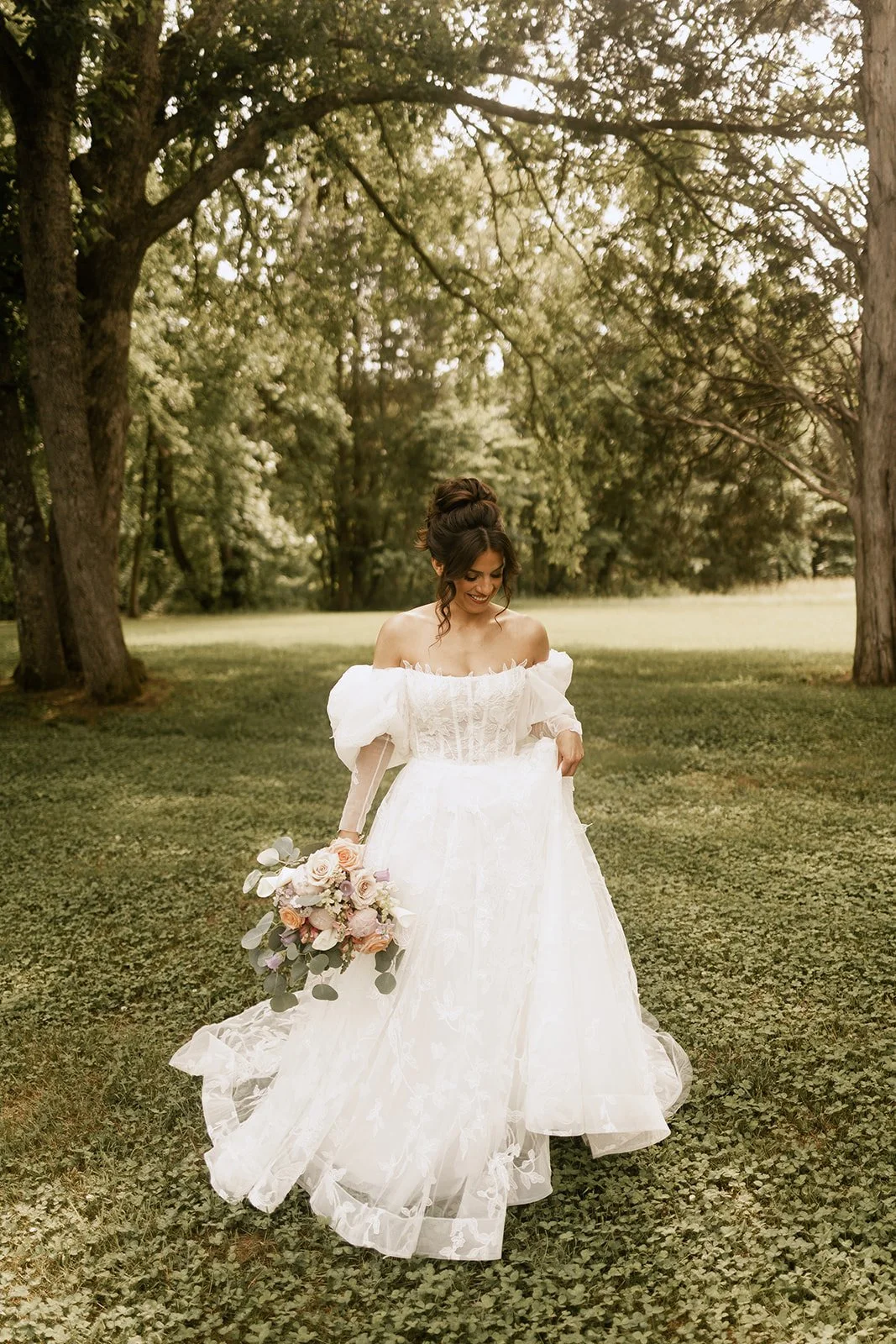 A bride in a white wedding dress standing on grass and holding a floral bouquet in an outdoor park setting.