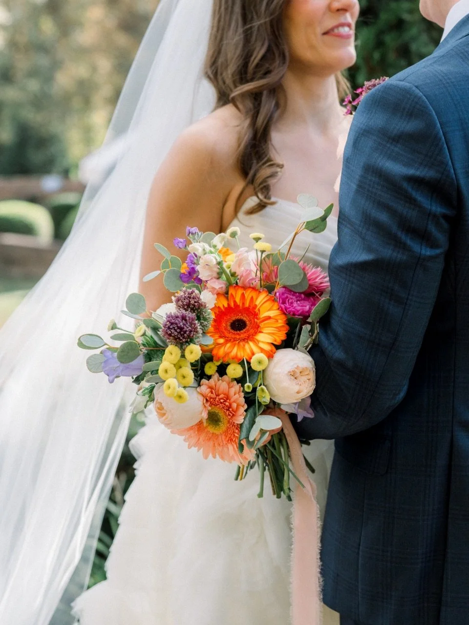 Bride holding a colorful bouquet of flowers, standing close to groom outdoors.
