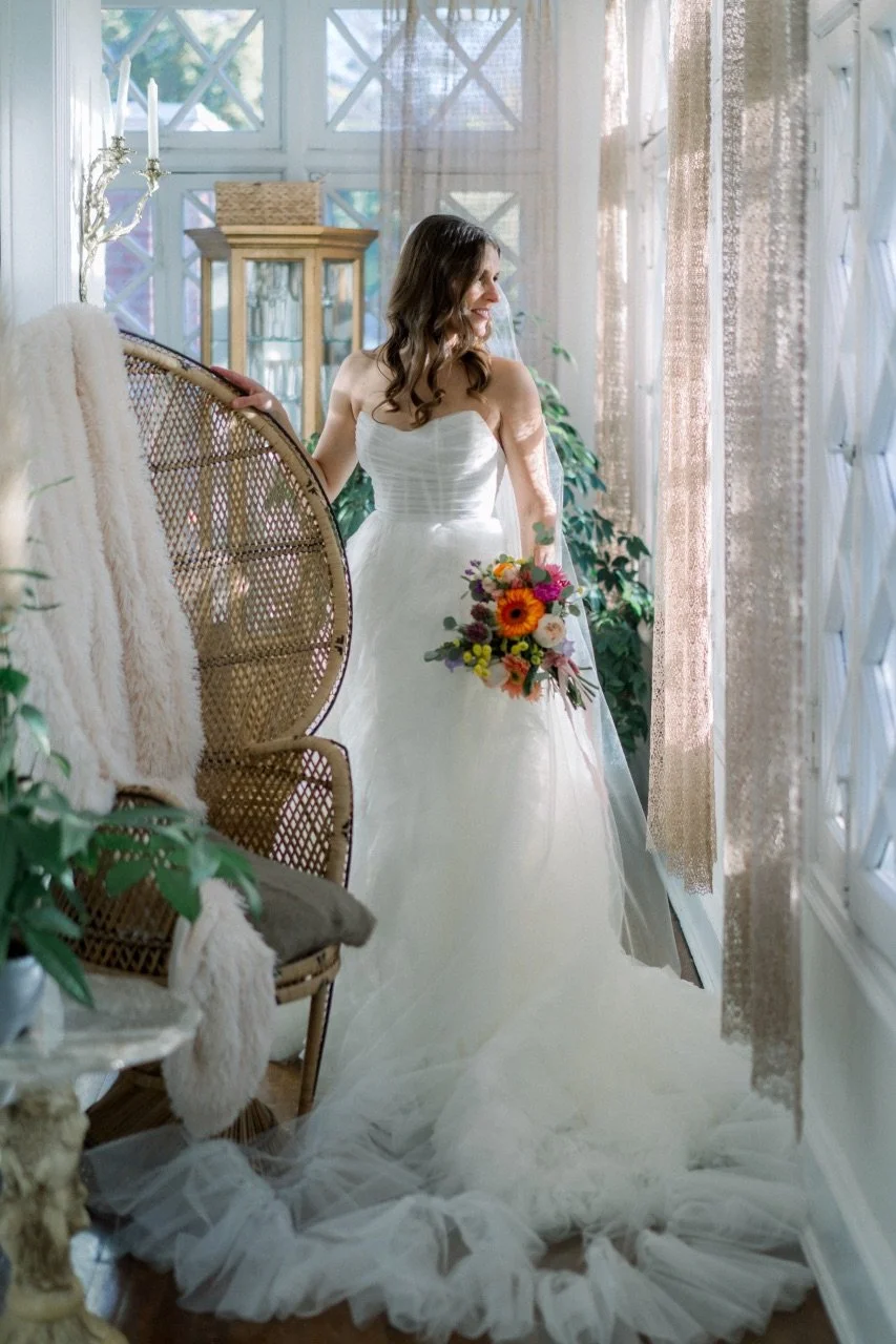 A bride in a white wedding dress holding a colorful bouquet, smiling and standing near large windows with curtains, surrounded by greenery and vintage furniture in a bright, airy room.