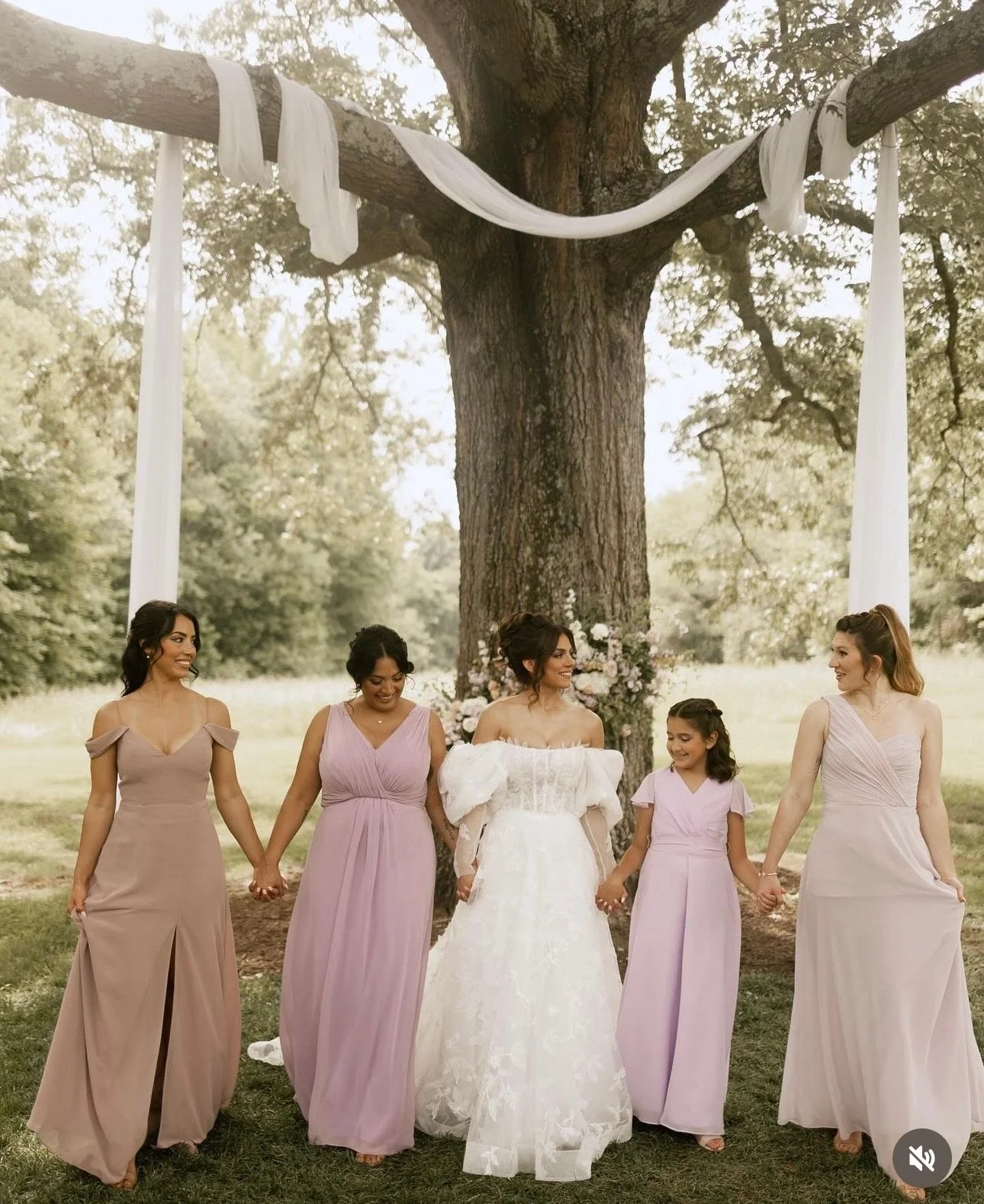 Group of women in wedding party holding hands, standing under a large tree with white drapery and floral arrangement, outdoor setting.