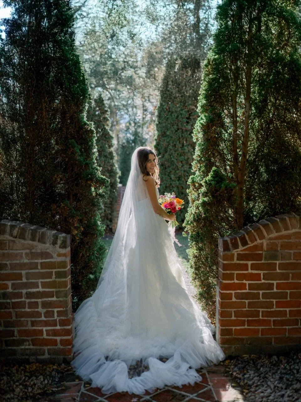 A bride in a white wedding gown holding a colorful bouquet, standing in a garden framed by greenery and a brick archway.