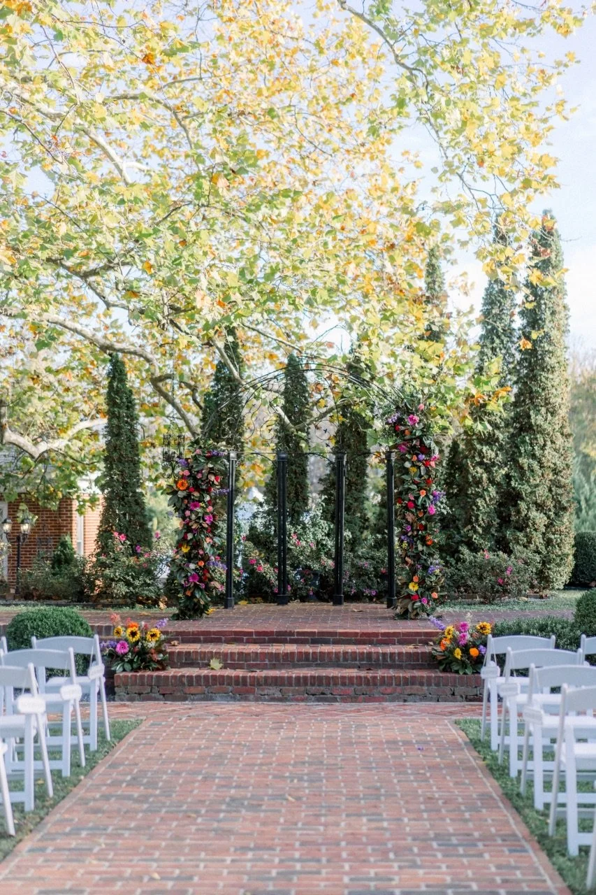 Wedding altar decorated with flowers on black metal arches, set outdoors on brick stairs, with white chairs arranged on either side on a brick pathway, surrounded by trees with yellow leaves.