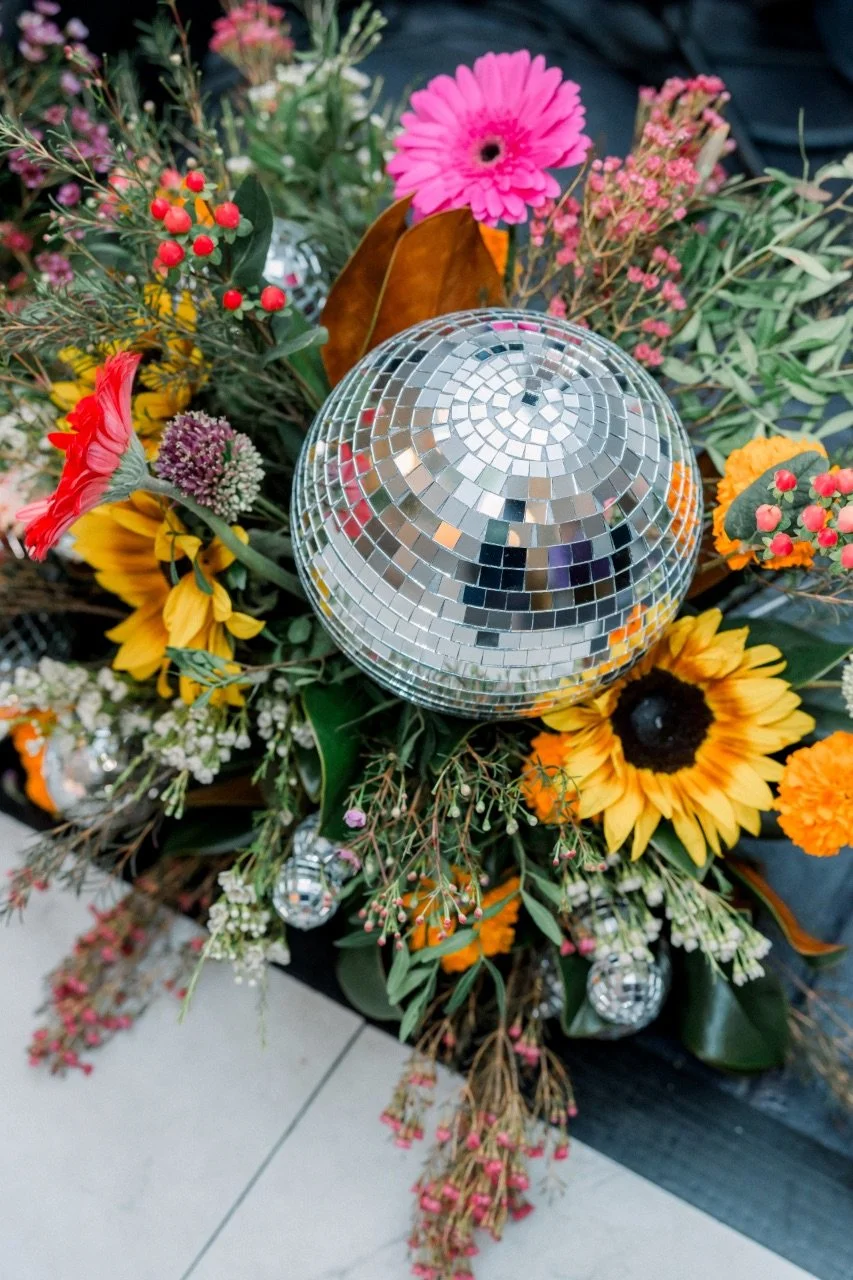 A colorful flower arrangement with sunflowers, pink zinnias, berries, and greenery, topped with a reflective disco ball.