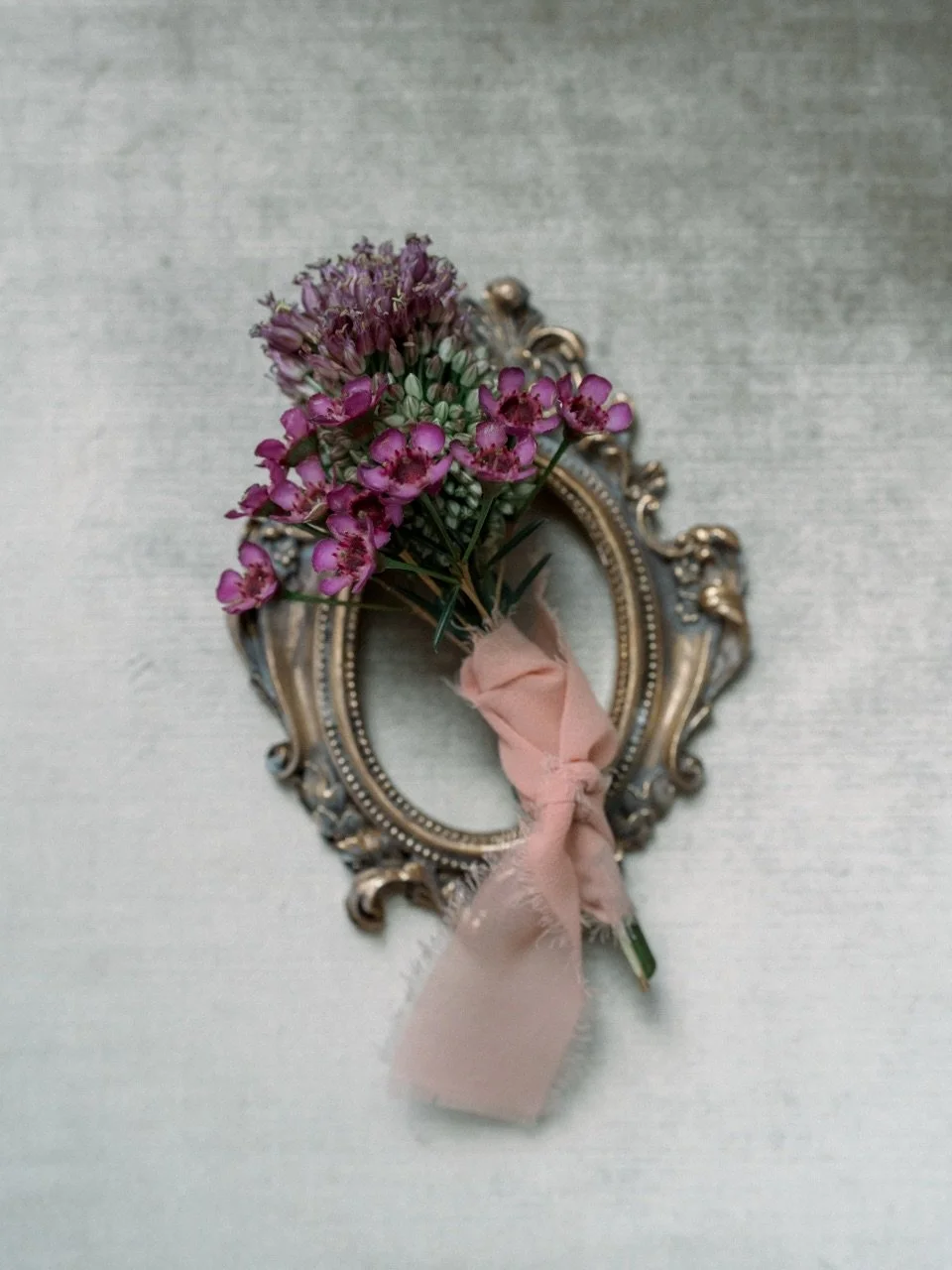 A small bouquet of purple flowers with green stems tied with pink ribbon, placed inside an ornate, vintage-style gold and silver picture frame on a light-colored textured surface.