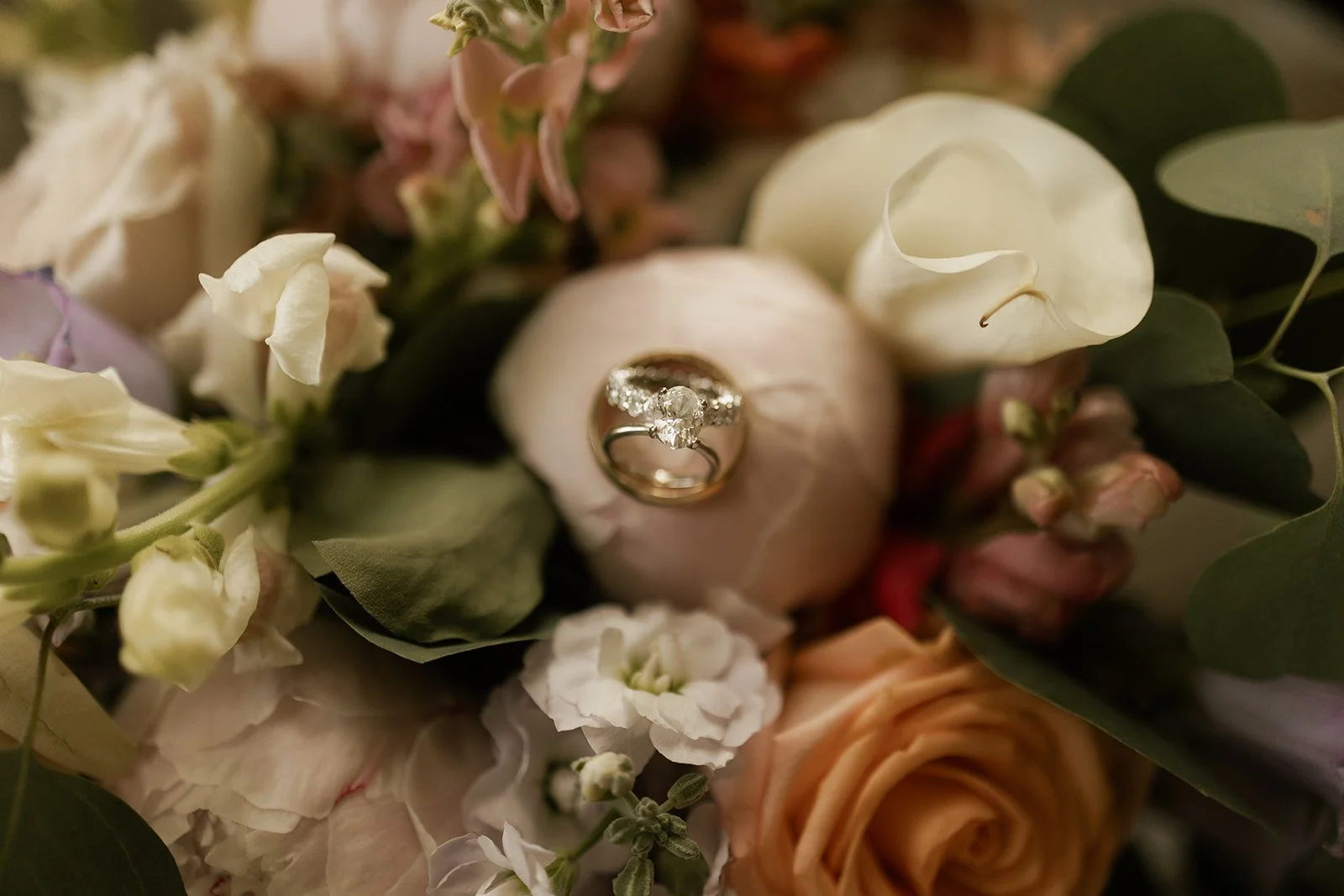 Close-up of a wedding ring with diamonds on a flower among various pastel-colored flowers and greenery.
