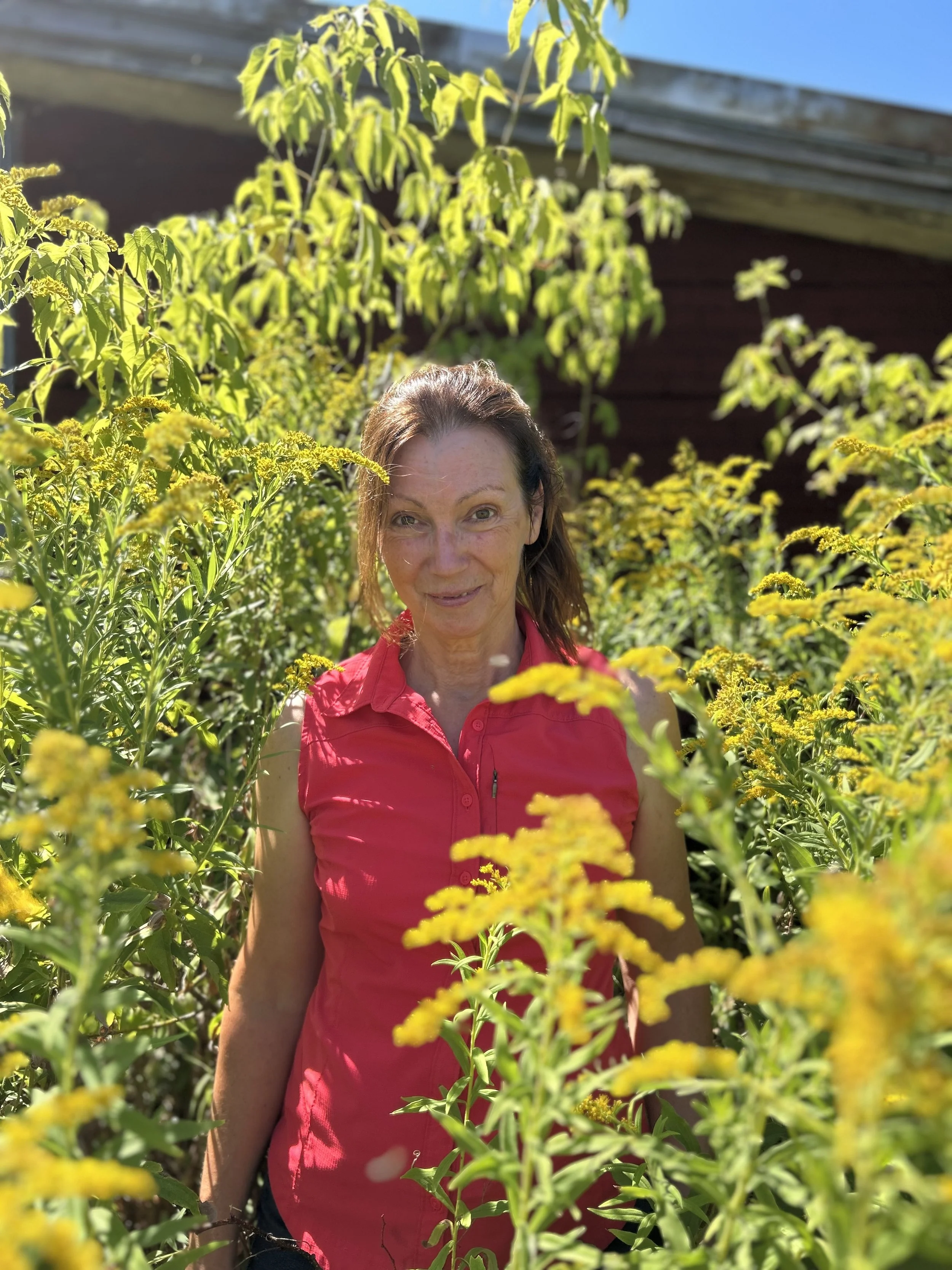 Femme dans un jardin entourée de fleurs jaunes et de végétation verte, portant un vêtement rouge, sous un ciel bleu.