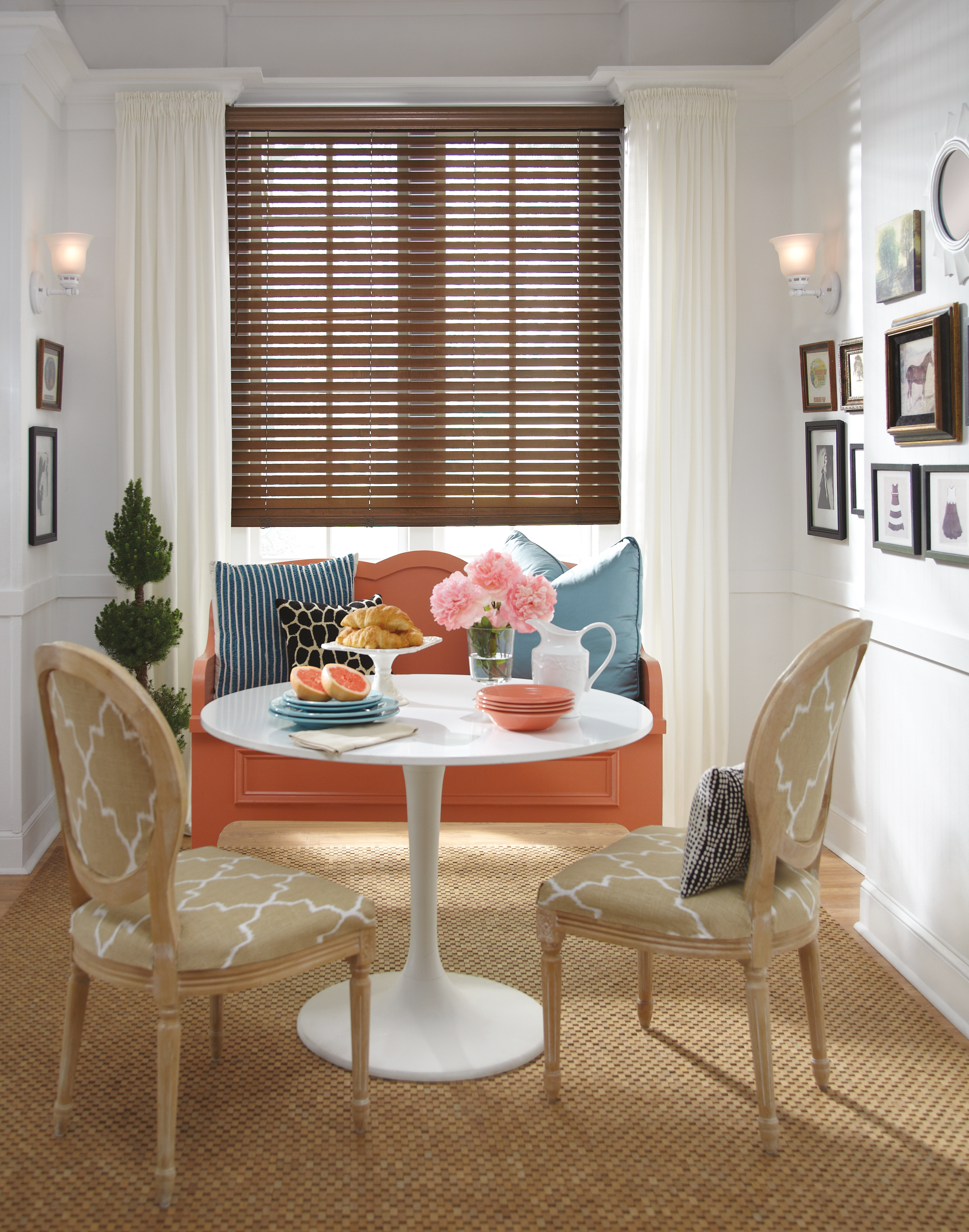 A cozy dining nook with a round white table set with pink bowls, a pitcher, a vase of pink flowers, and pastries. There are two beige patterned chairs around the table and a coral-colored bench with striped and solid pillows by a window with white cu