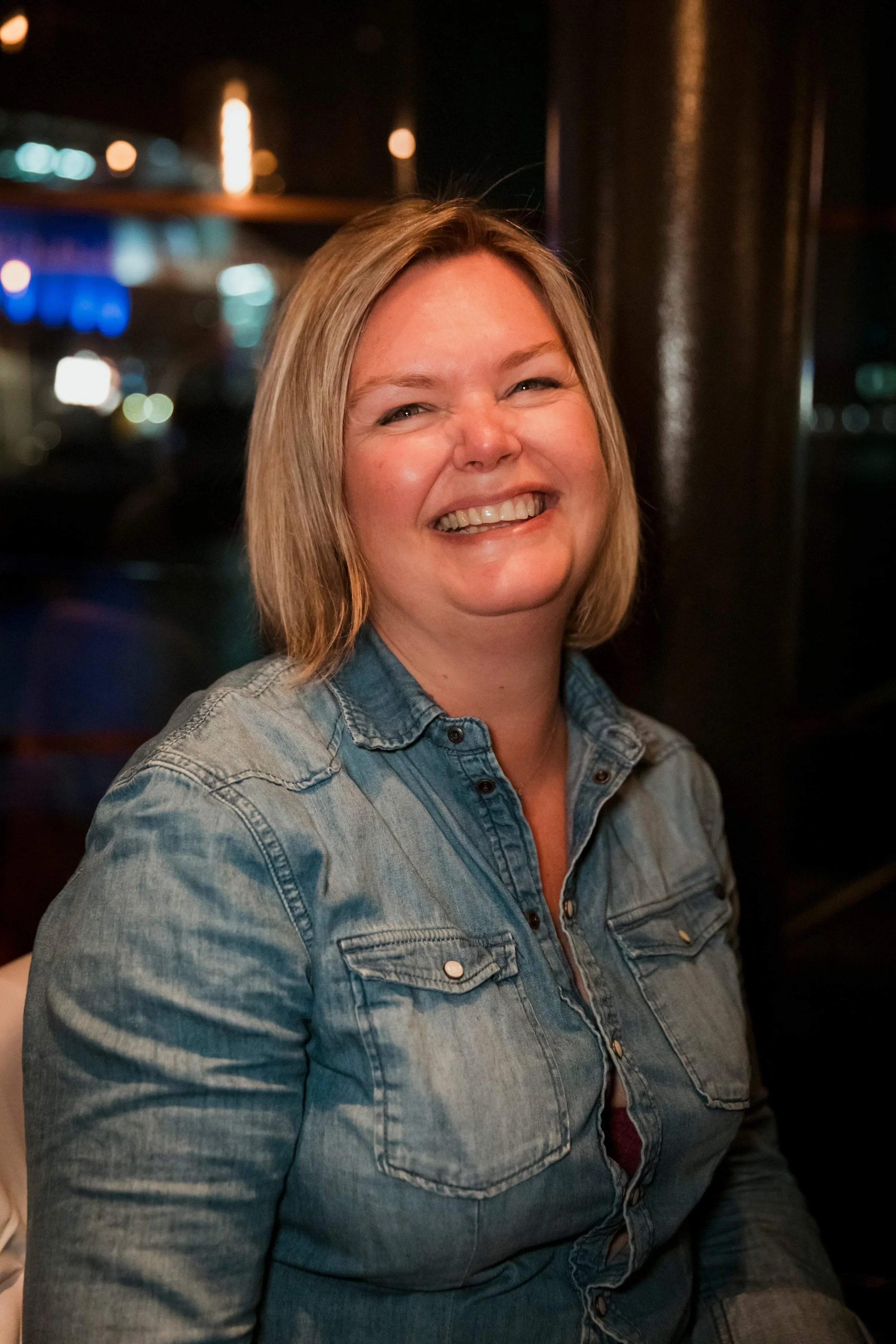 A woman with shoulder-length blonde hair smiling and wearing a denim shirt, in a dimly lit setting with blurred colorful lights in the background.
