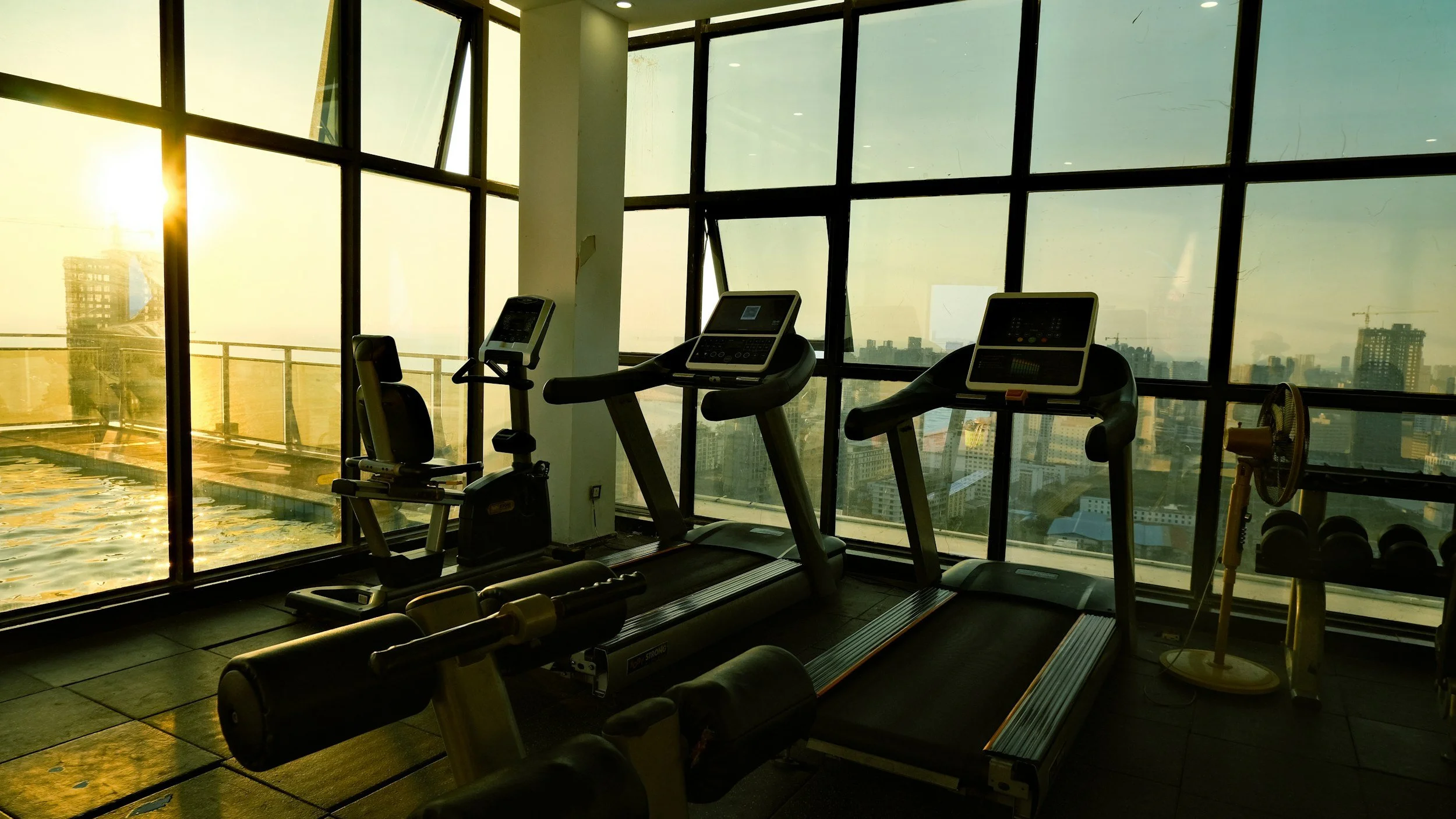 Empty gym with treadmill, stationary bike, and elliptical in front of floor-to-ceiling windows showing city skyline at sunset.