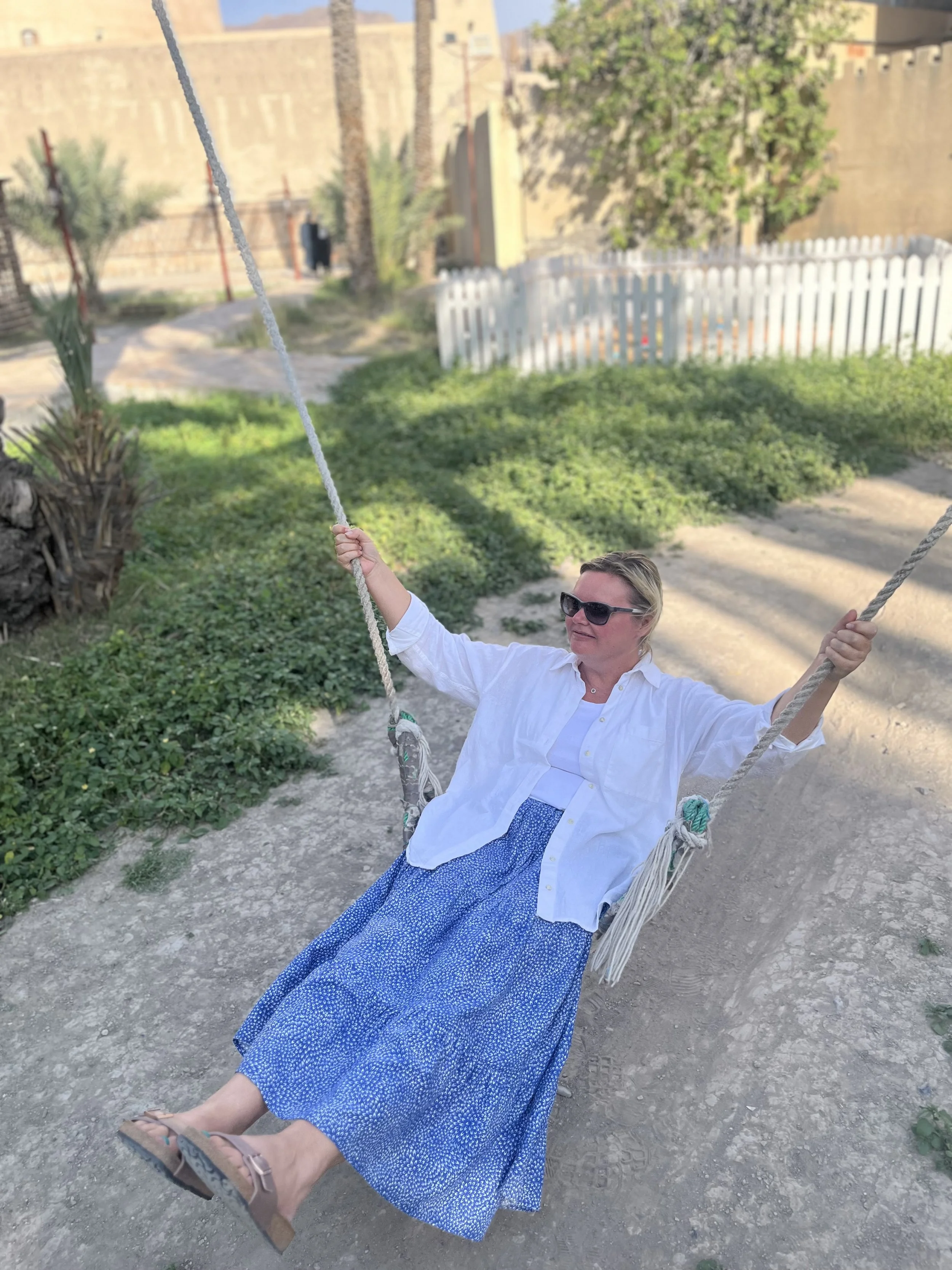 Woman in sunglasses and a white shirt sitting on a swing outdoors with greenery and a white picket fence in the background.