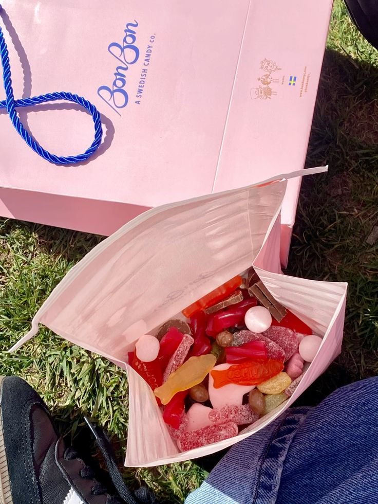Open pink gift bag containing various colorful gummy candies on grass, with a pink BonBon box beside it.