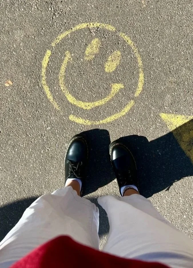 A person's feet in black shoes and white pants standing on asphalt with a yellow smiley face drawn on the ground.