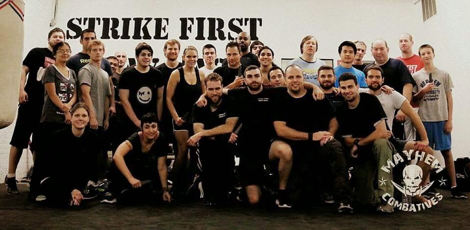 Group of people, including both men and women, standing together for a group photo in a gym or fitness facility. The background features a large banner that says "STRIKE FIRST" and a logo for "Mayhem Combatives" with a skull symbol.