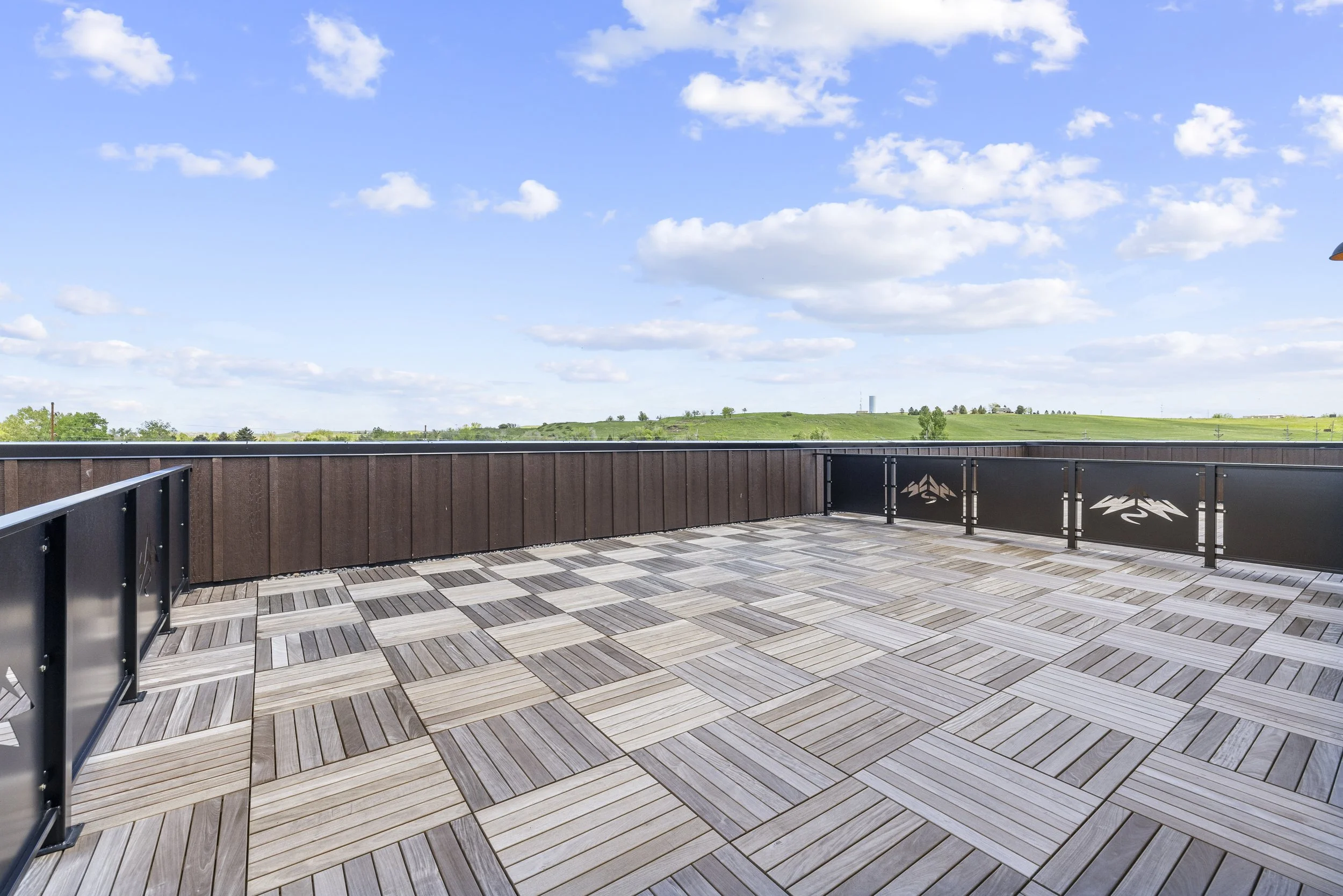 Empty rooftop deck with light-colored wooden tiles, metal railings, and a scenic view of green hills and a blue sky with scattered clouds.