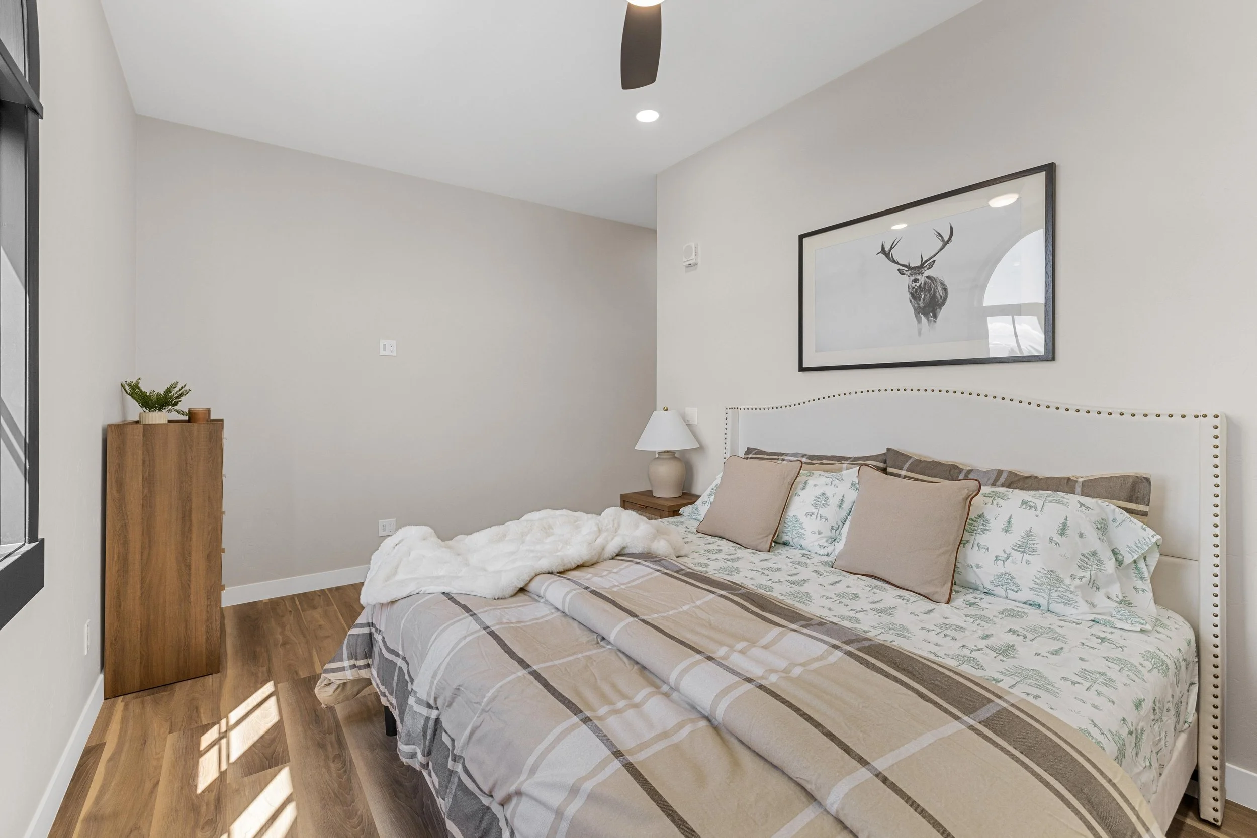 Modern bedroom with bed covered in plaid and woodland-themed bedding, framed deer print on wall, wooden side table with lamp, small plant on tall wooden dresser, and hardwood flooring.