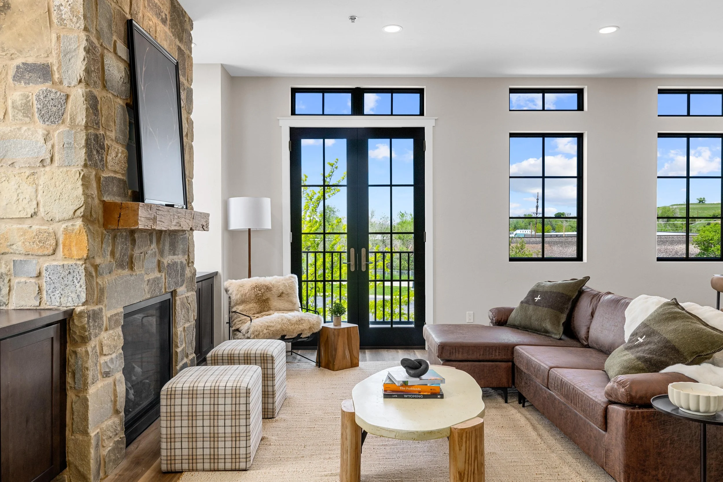 Living room with stone fireplace, wall-mounted TV, black framed glass door leading outside, multiple windows showing blue sky and greenery, brown sectional sofa with pillows, fur-covered chair with ottomans, round coffee table with books and decorati