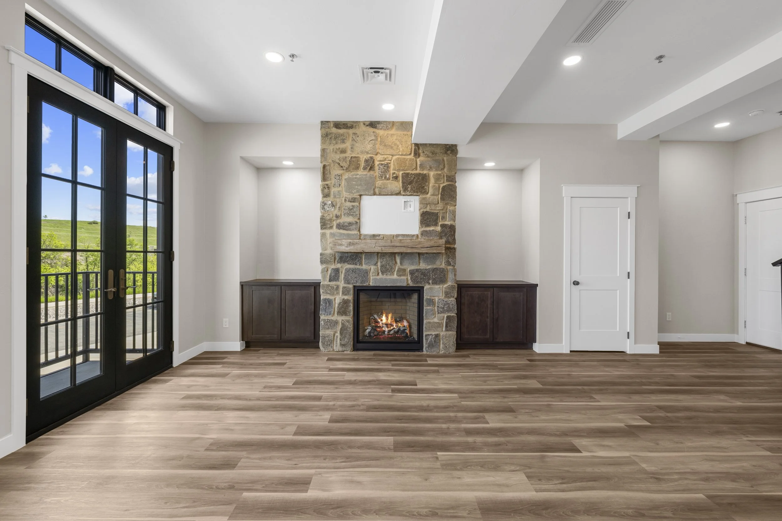 Empty living room featuring a stone fireplace, dark wood cabinets, white walls, hardwood floors, and black-framed glass sliding doors leading to an outdoor patio with a view of green fields and blue sky.