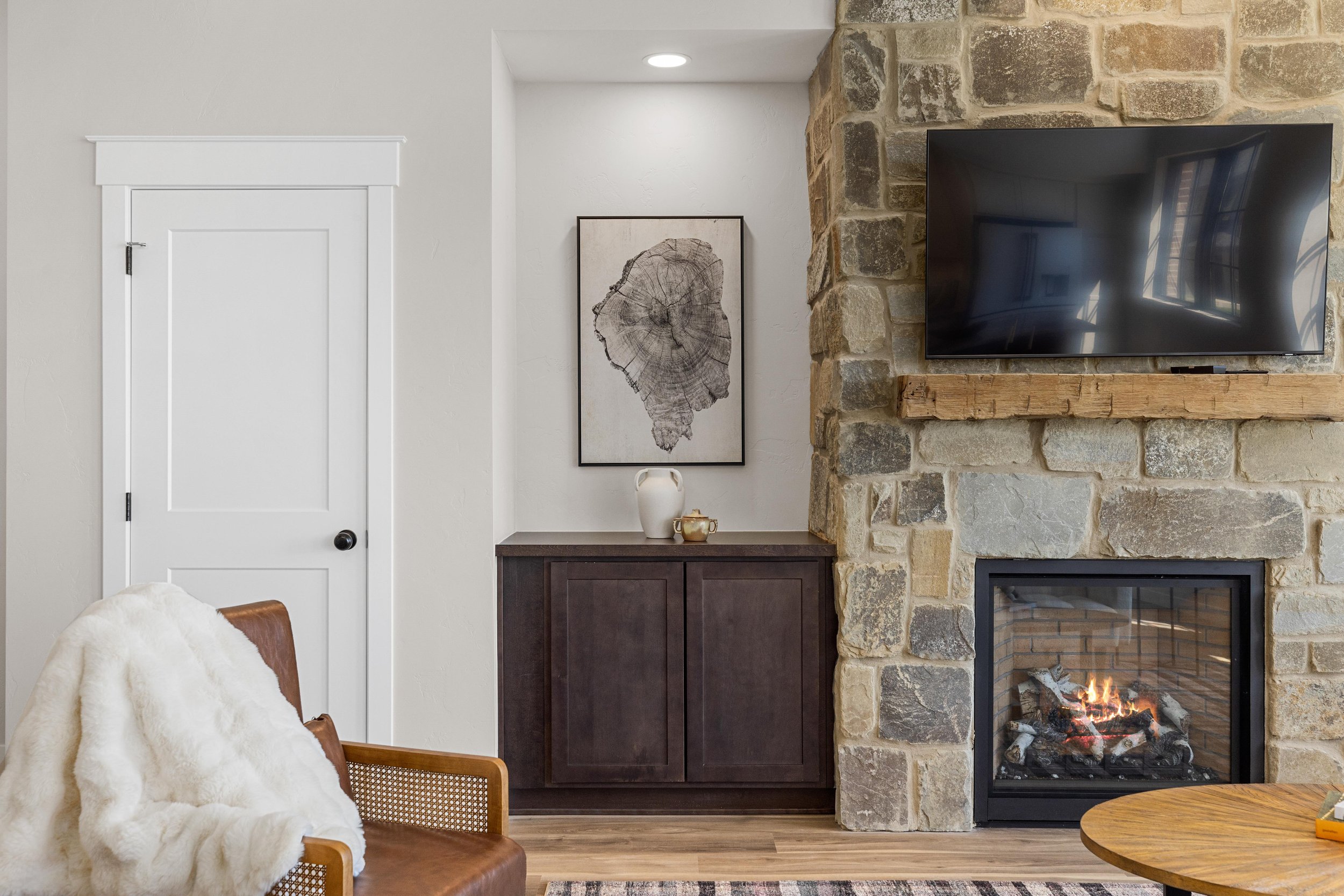 Living room with a stone fireplace, wall-mounted TV, dark wood cabinet, and framed artwork of a tree trunk cross-section. There is a wooden armchair with a white blanket and a round wooden table.
