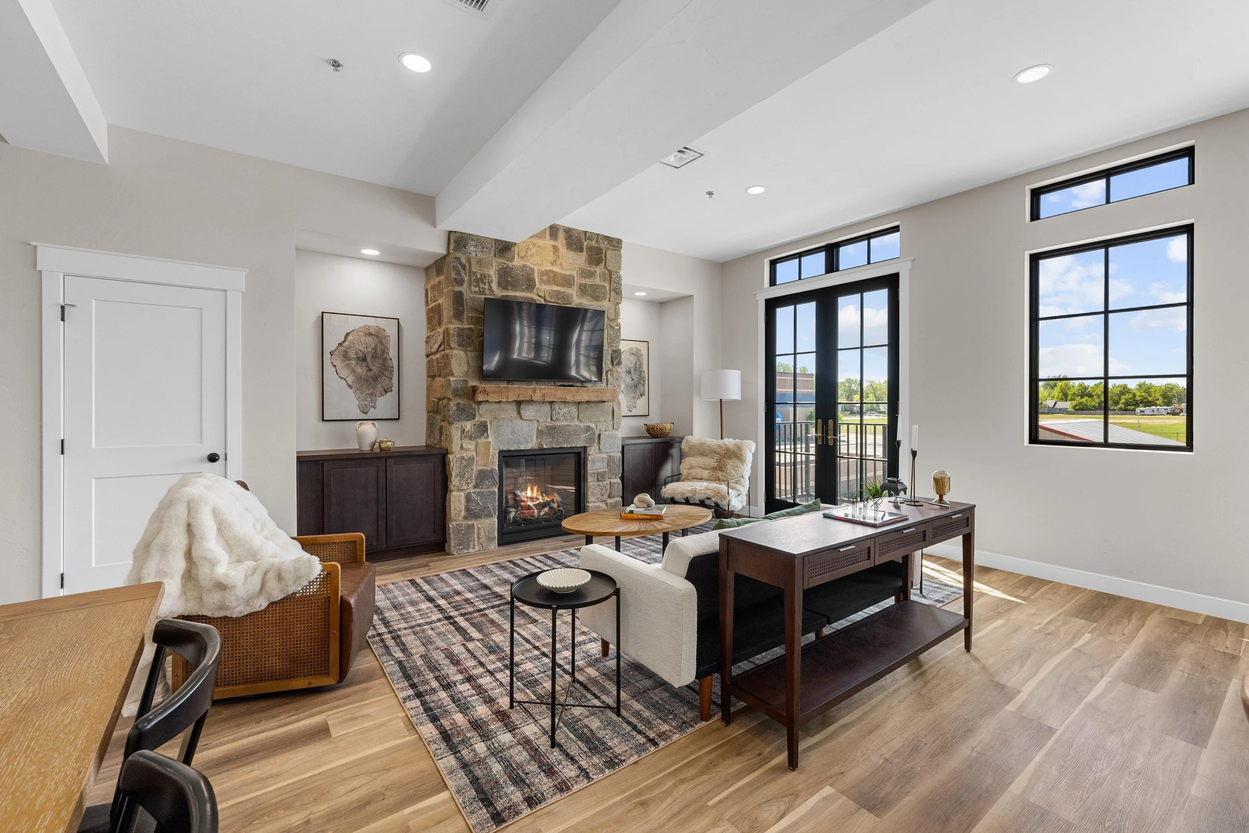 Living room with a stone fireplace, a TV above it, a sliding glass door, and large windows showing an outdoor view. The room features modern furniture, including a white sofa, wooden coffee table, and patterned area rug.