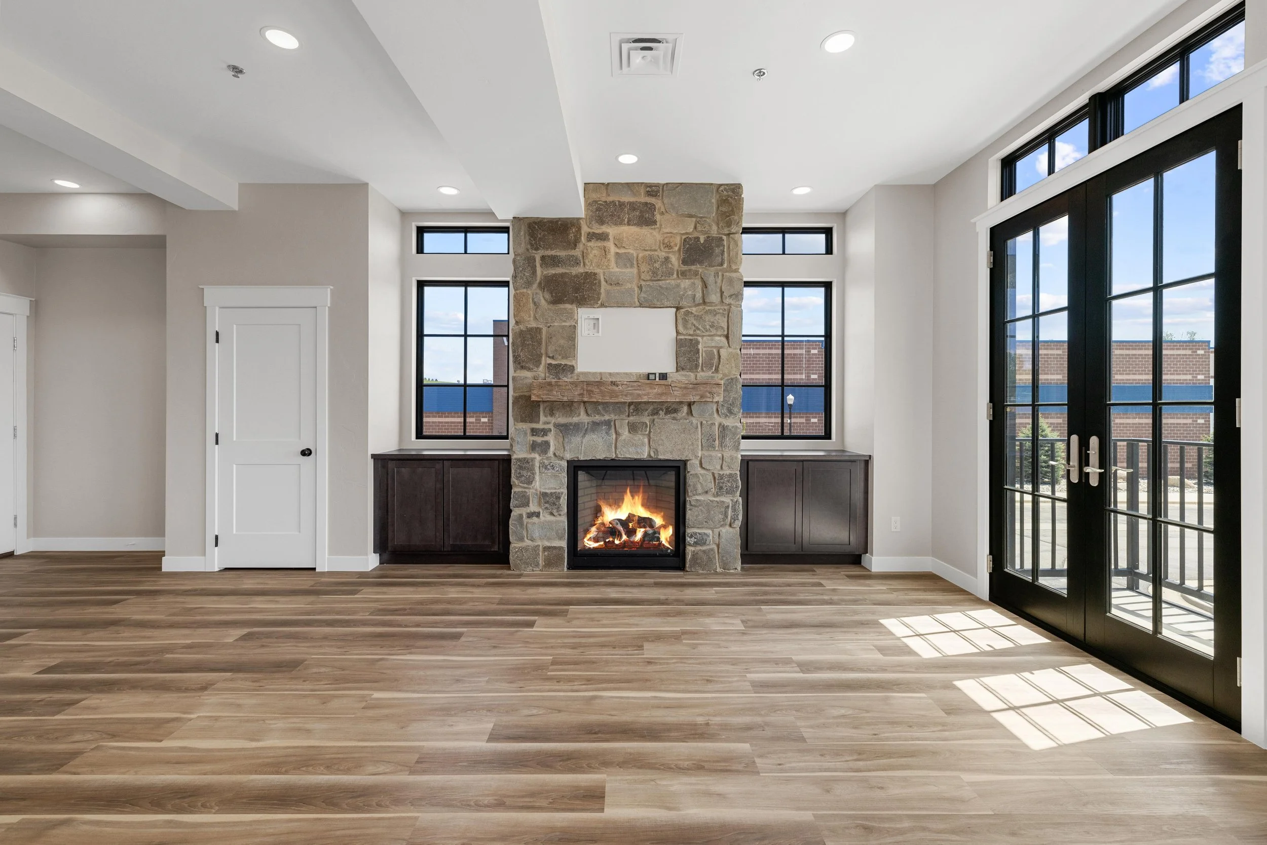 A spacious living room in an Ice Haus Condo for sale in Sheridan, Wyoming, with a stone fireplace, wooden floors, and large windows and glass doors providing natural light.