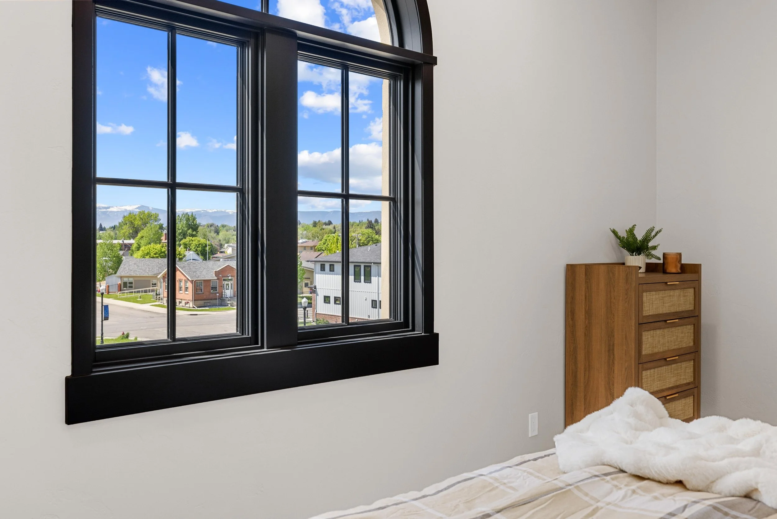 A bedroom in an Ice Haus Condo for sale in Sheridan, Wyoming,  with a large black-framed window showing a view of houses, greenery, and distant mountains under a blue sky with clouds.