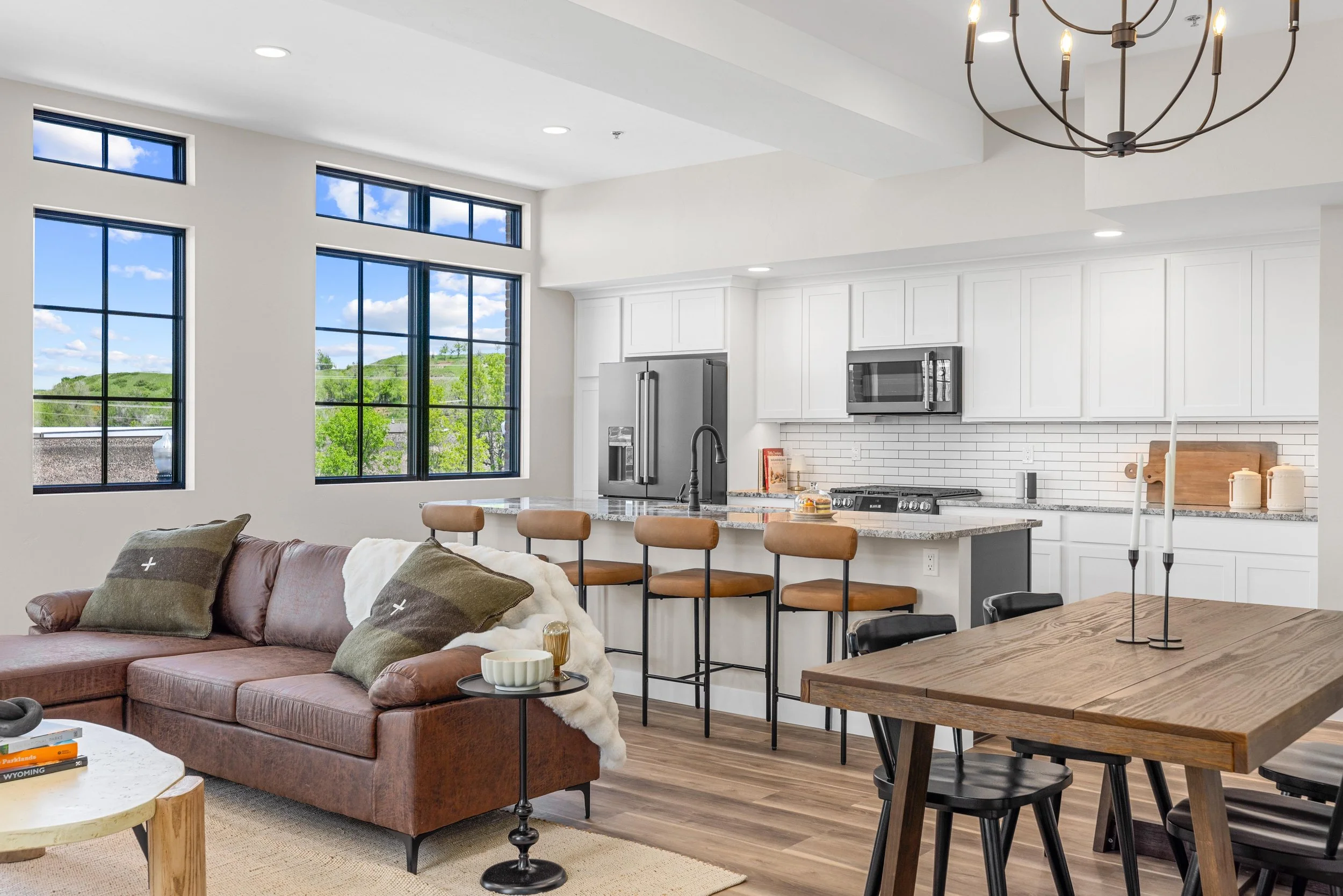 Open-concept living room and kitchen with large windows, white cabinetry, stainless steel appliances, brown leather couch, black table, and wooden bar stools.