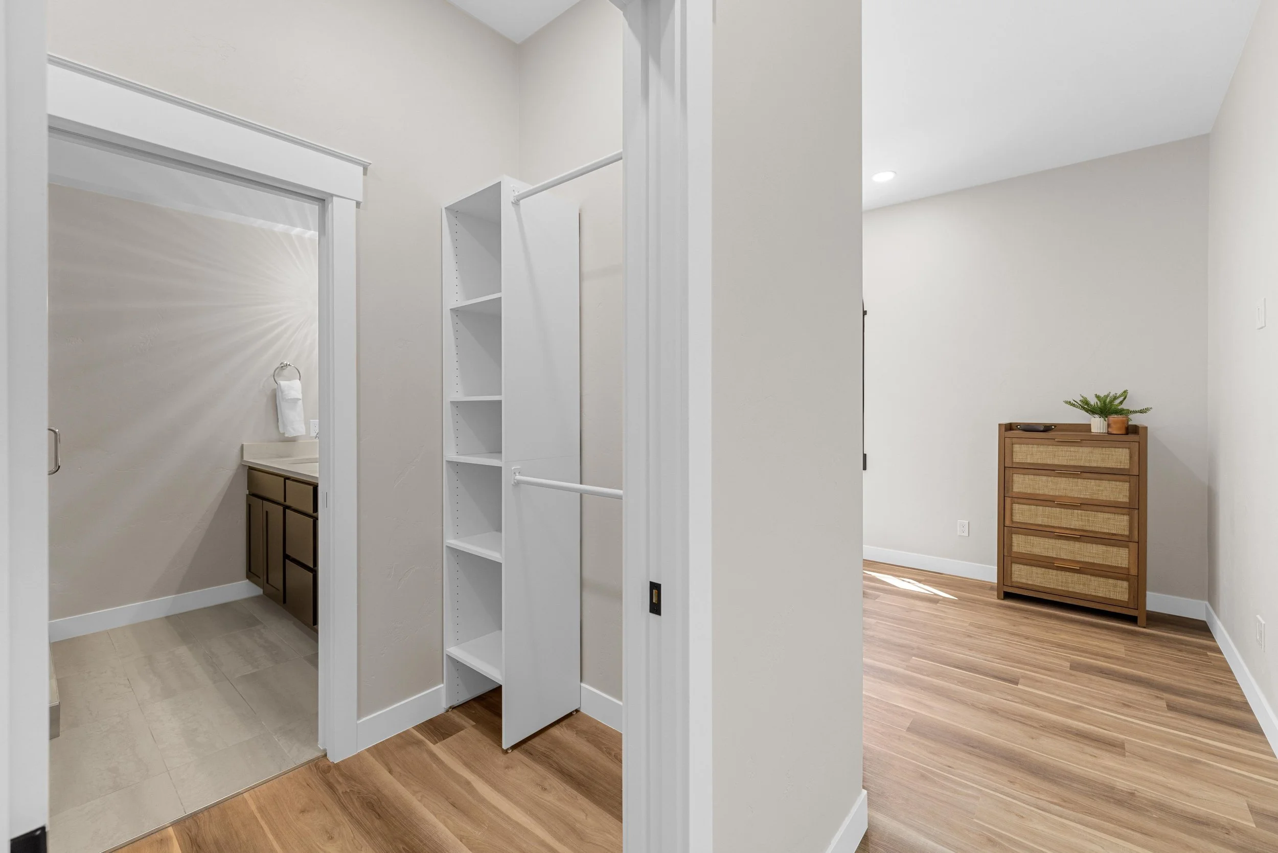 A room with a closet and a bathroom leading into the bedroom, featuring wooden flooring and white walls, with a small plant on a wooden dresser, in an Ice Haus Condo for sale in Sheridan, Wyoming.