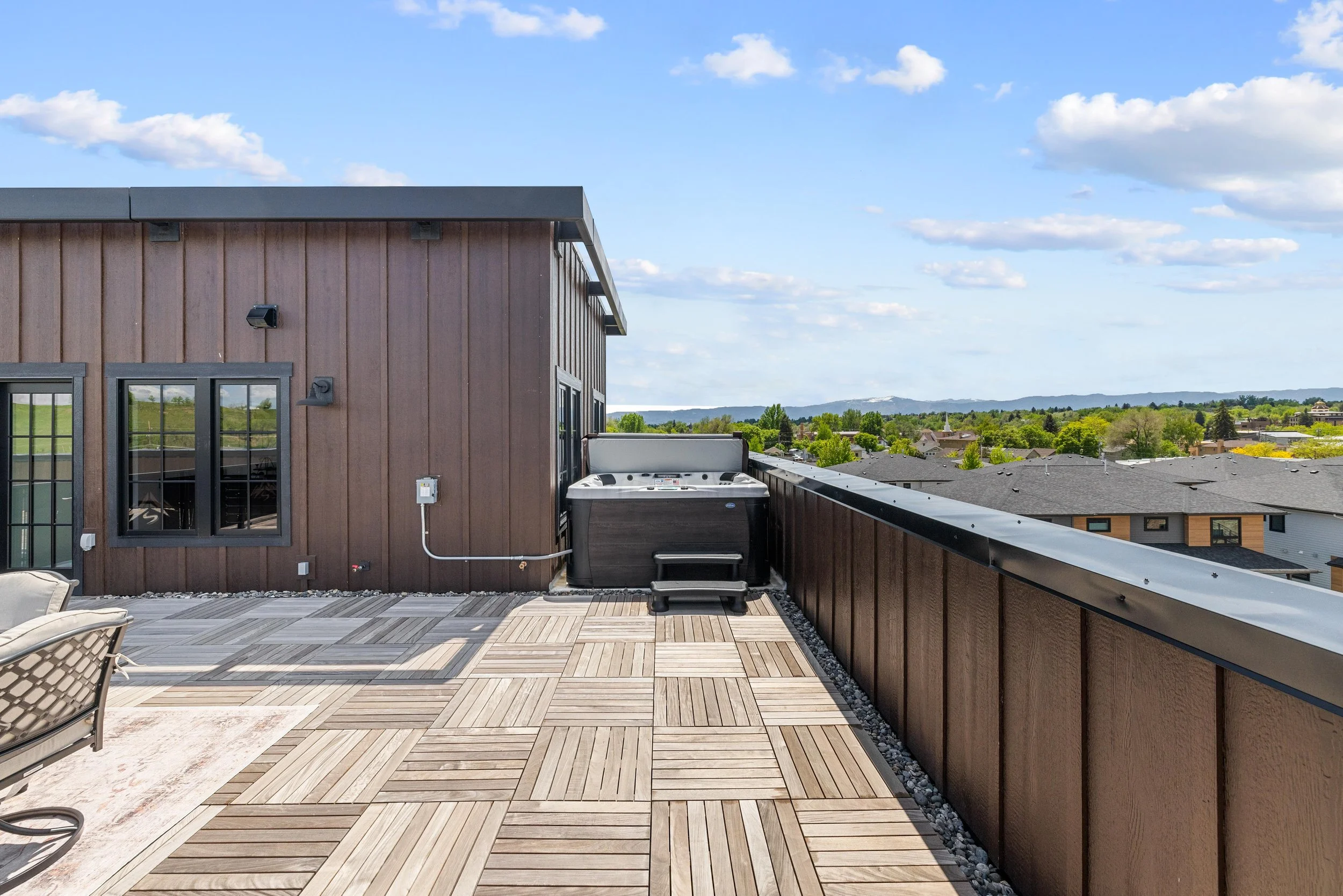 Rooftop balcony with wooden flooring, a hot tub, and view of neighboring houses and distant hills under a blue sky with clouds.