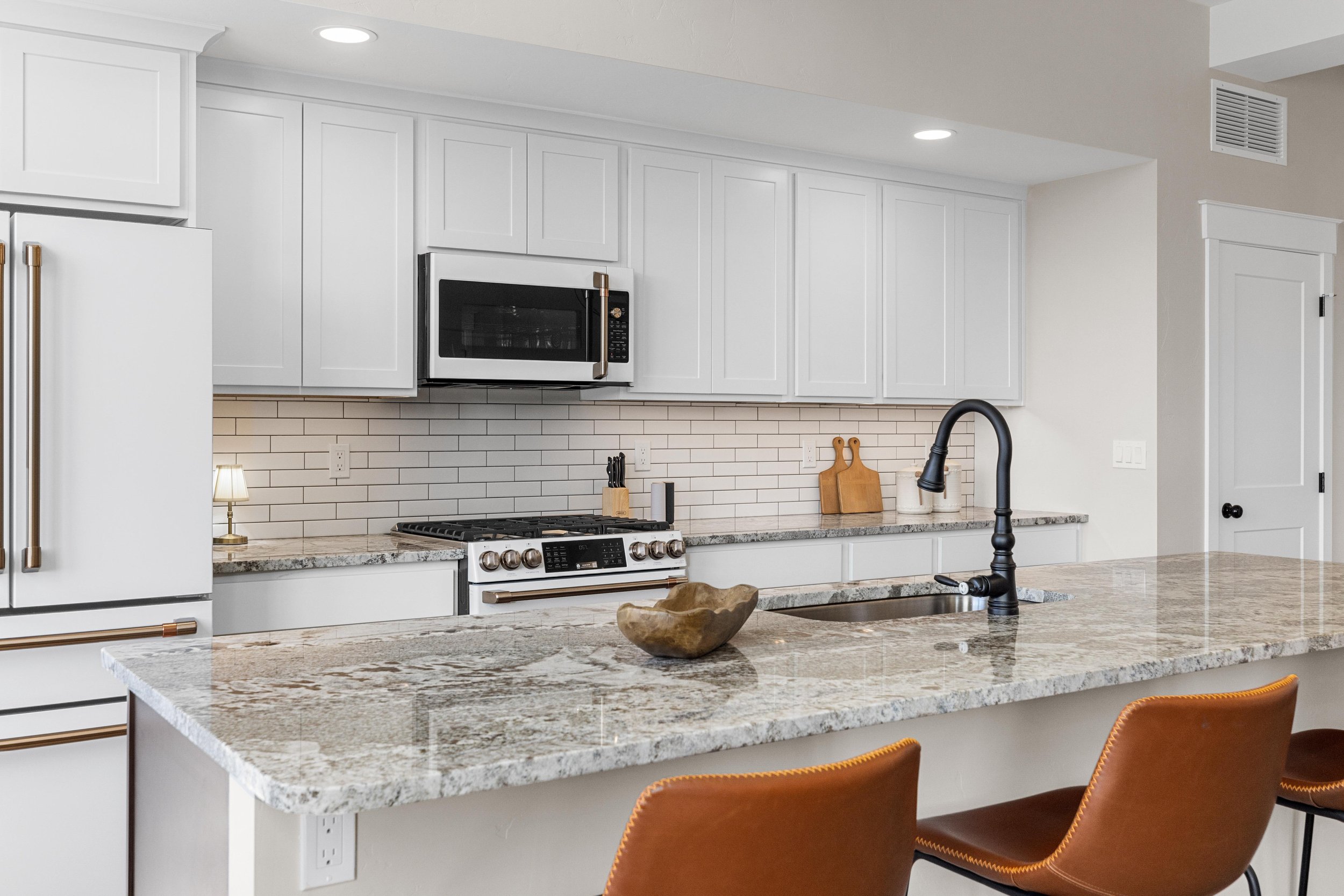 Modern kitchen with white cabinets, gray granite countertops, a black faucet, stainless steel appliances, and a white subway tile backsplash. There are three brown leather chairs in front of a kitchen island.