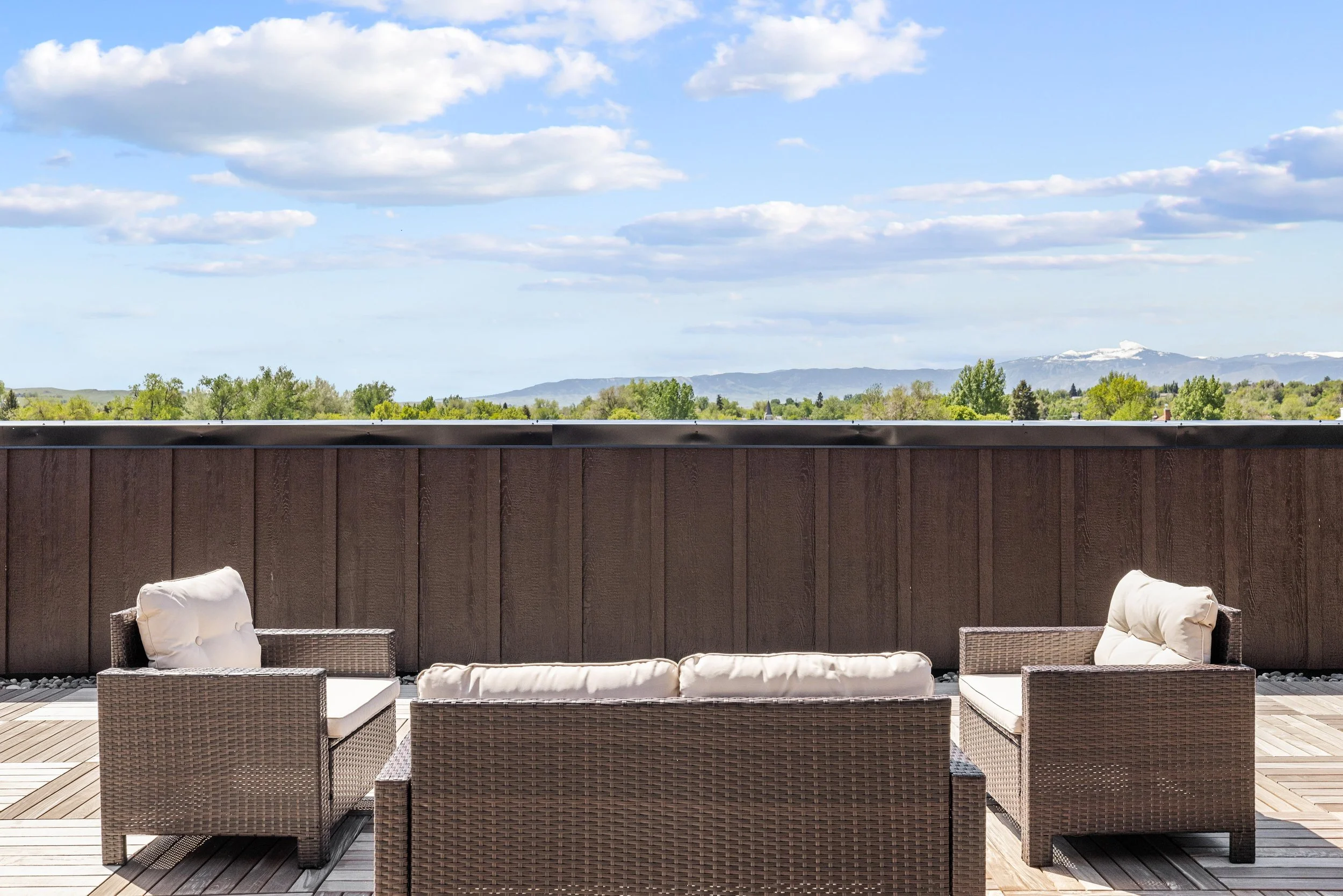 Outdoor patio with wicker furniture and white cushions, overlooking a landscape with green trees and distant mountains under a partly cloudy sky.