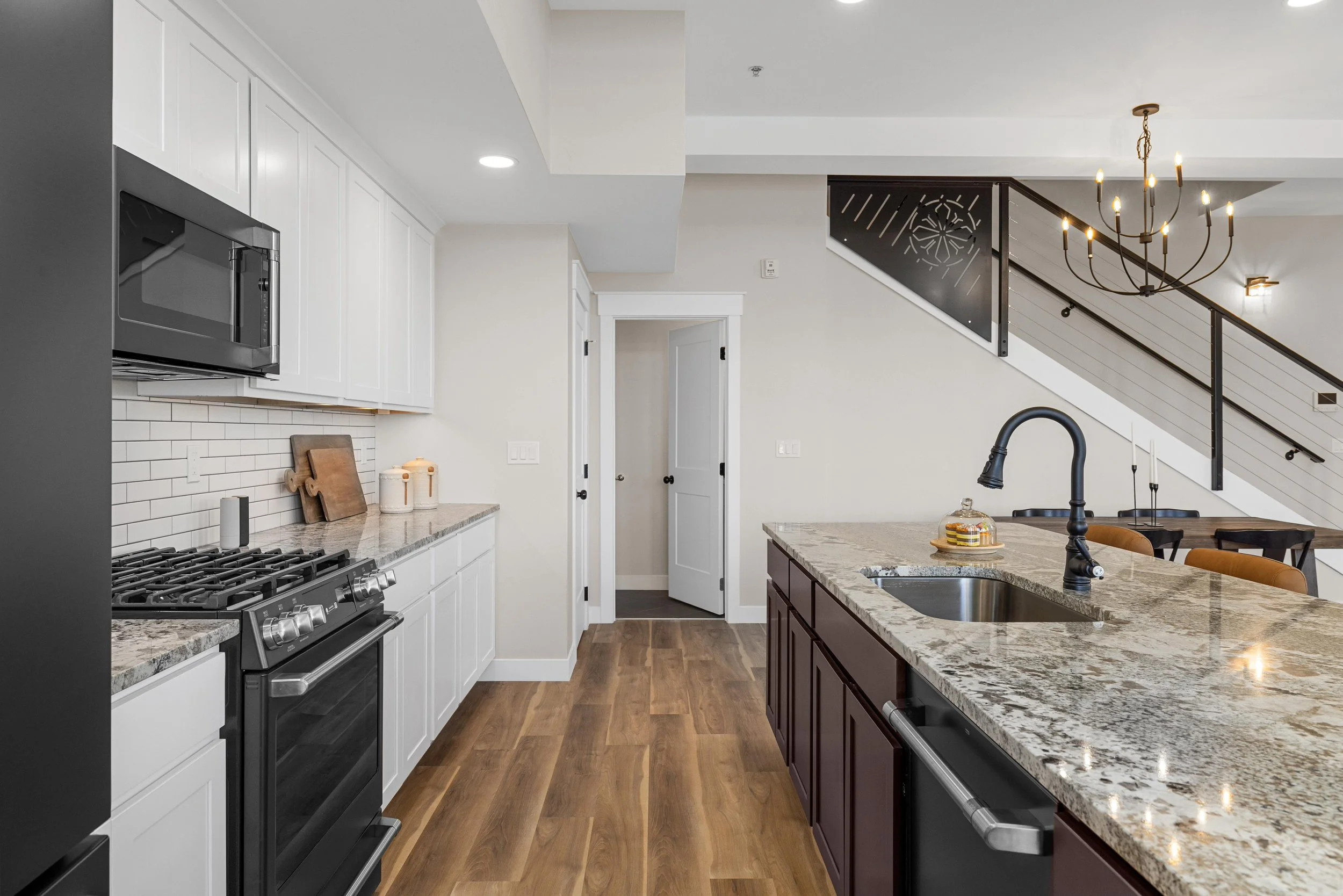 Modern kitchen with white upper cabinets, black and stainless appliances, granite countertops, a kitchen island with a sink, wooden floors, and a staircase with a chandelier overhead.