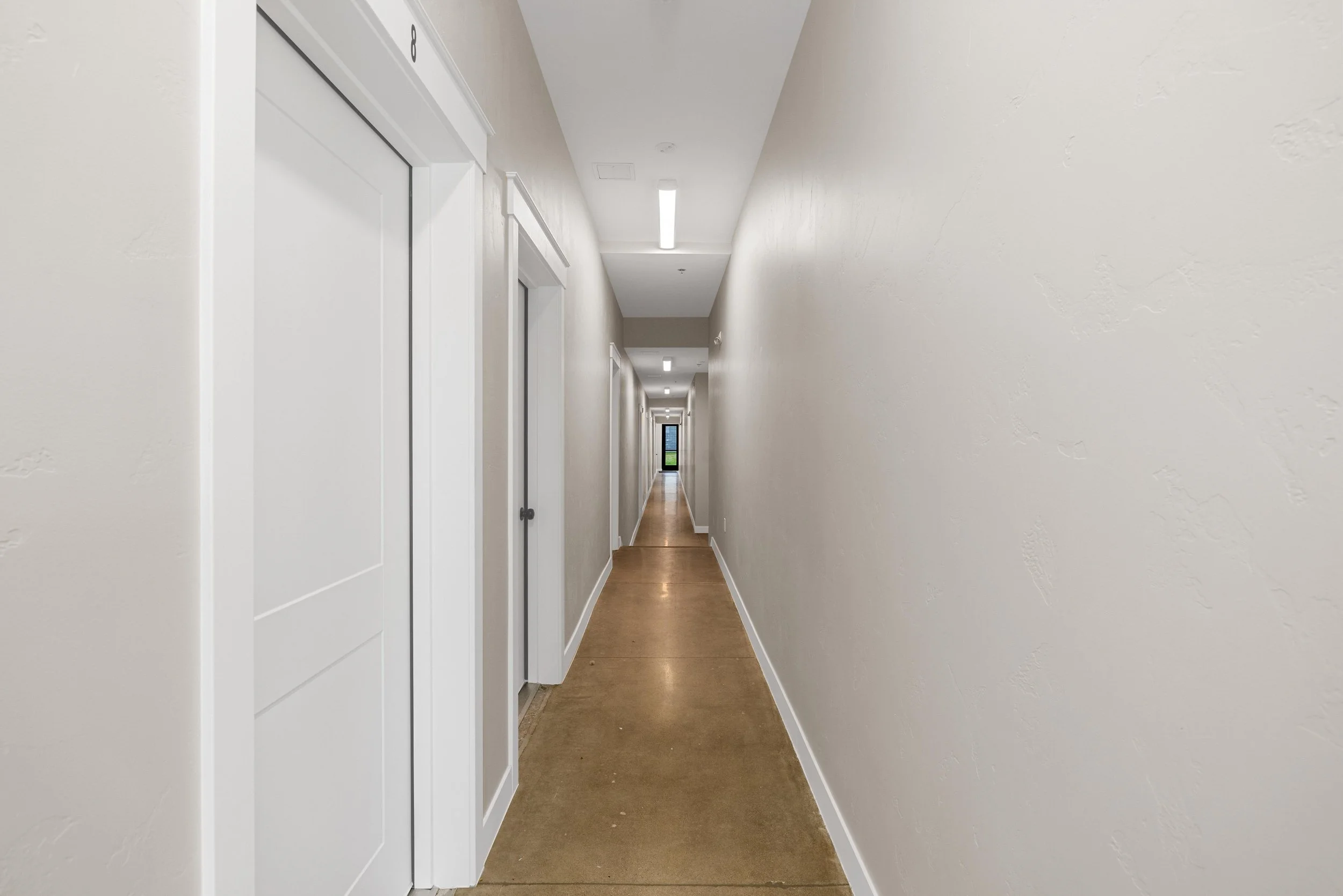 Long, empty hallway with white walls, closed white doors on the left, and a wooden floor, extending to a door with a window at the far end.