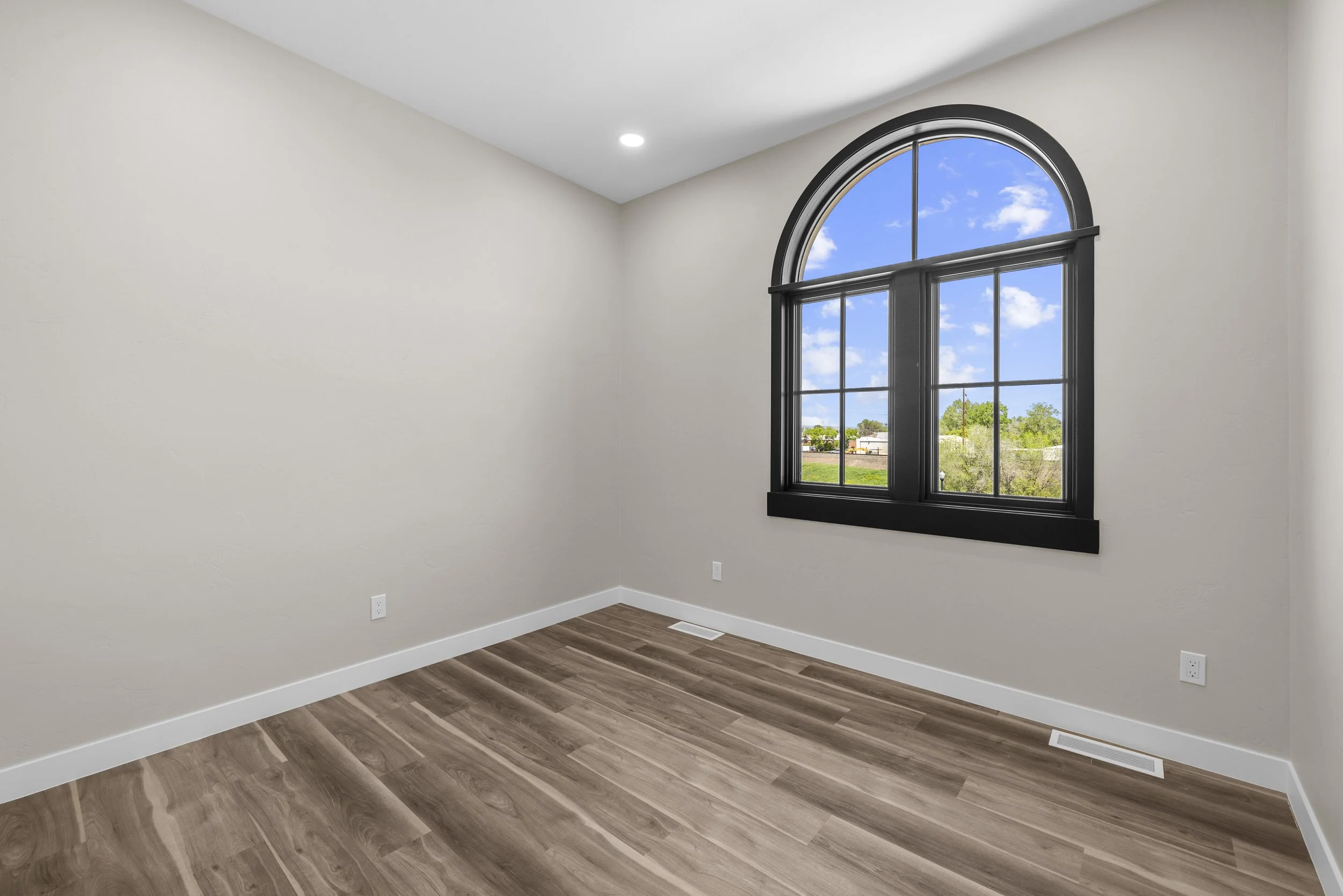 Empty room with beige walls, black-framed arched window showing a view of a sunny sky and green landscape, hardwood floor, and white baseboards.