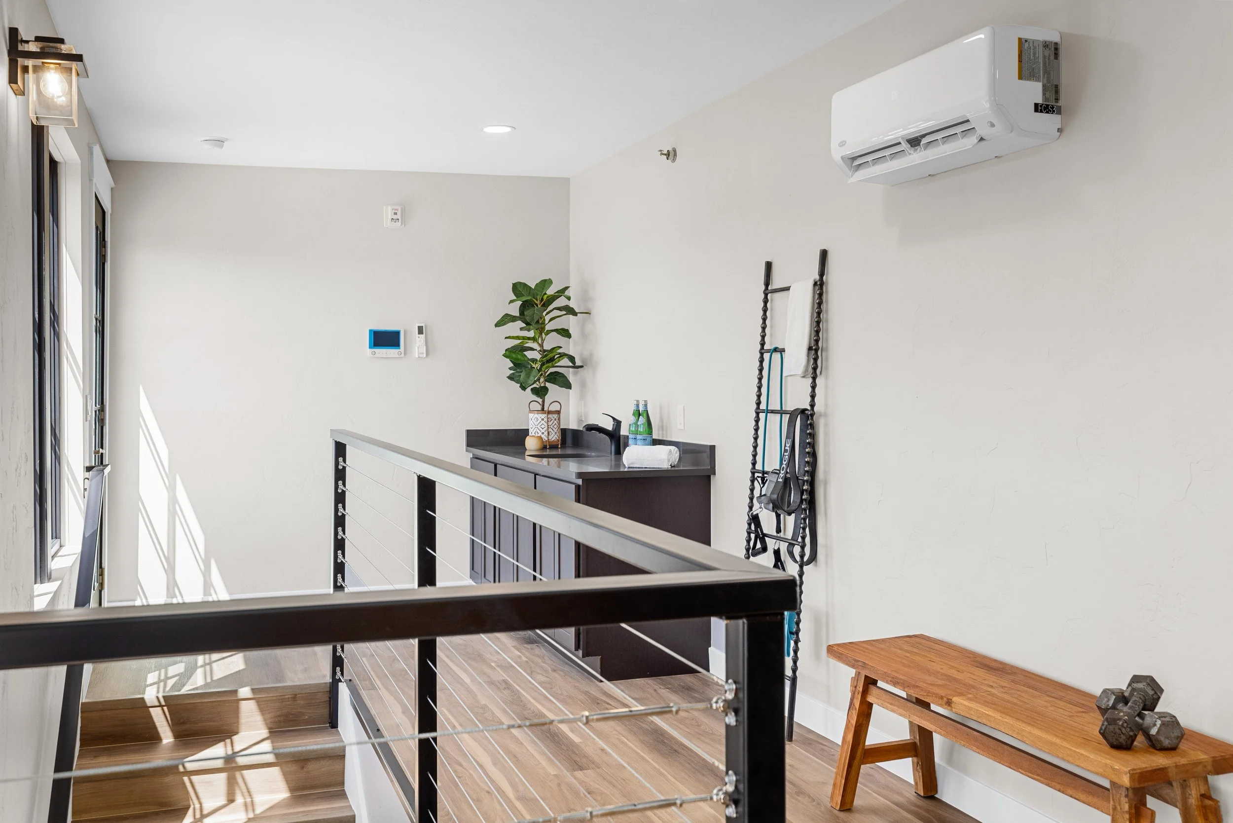 Interior of a modern home upstairs area with a black railing, wooden stairs, a small counter with a plant and two bottles of water, a towel, a ladder with exercise bands, a wooden bench, and dumbbells on it. An air conditioning unit is mounted on the