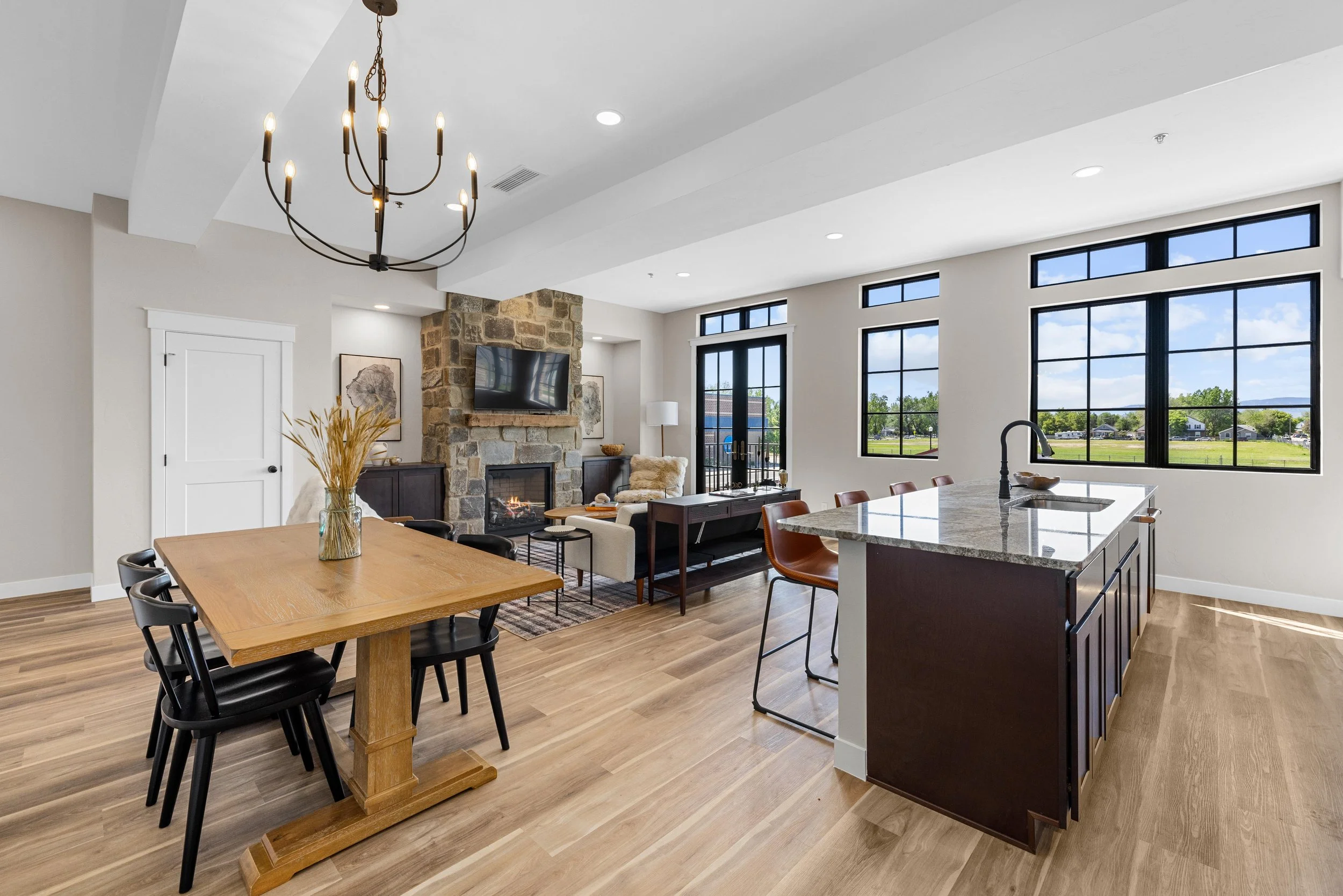 Open-concept living and dining area with large windows, a stone fireplace with a TV, a wooden dining table with black chairs, and a kitchen island with barstools.