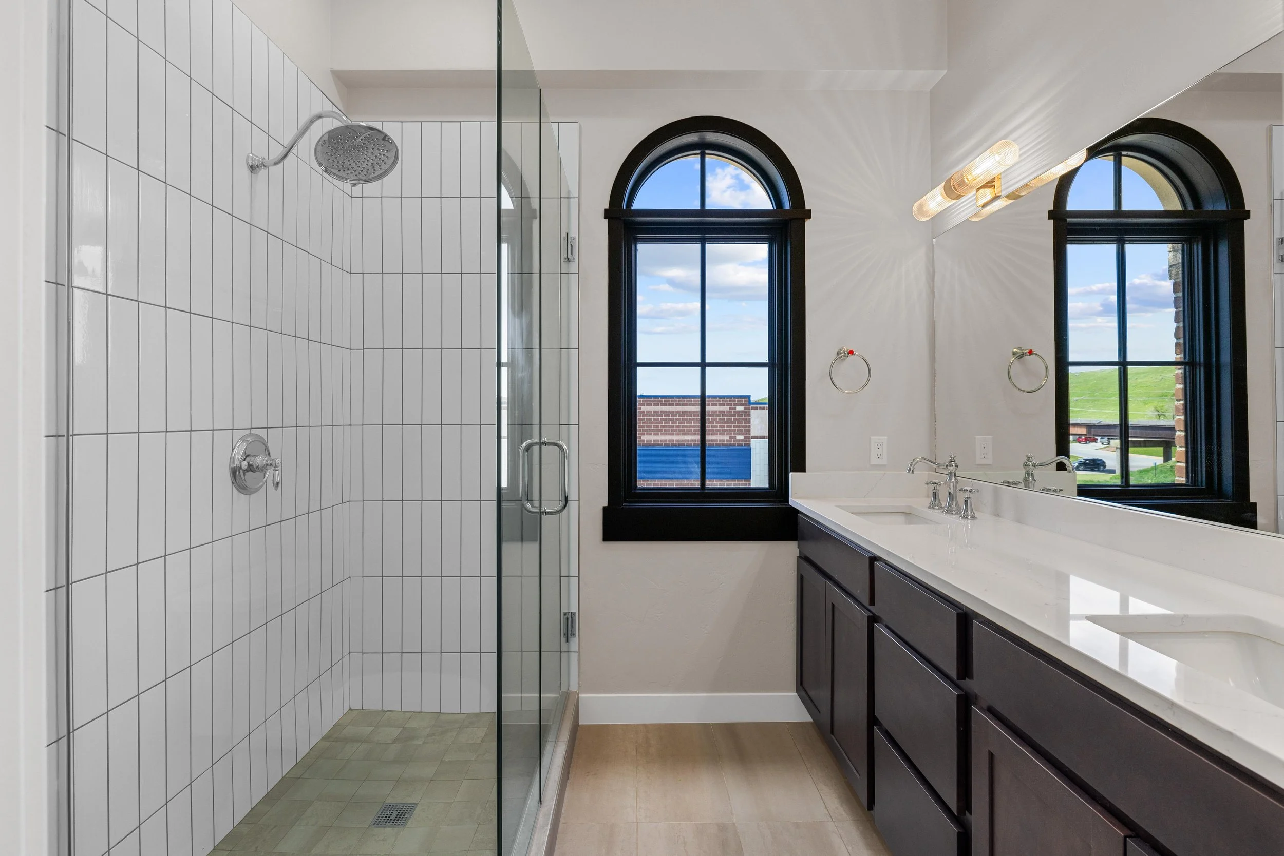 Bathroom with a glass-enclosed shower, a double sink vanity, large arched windows showing a view outside, and a large mirror.