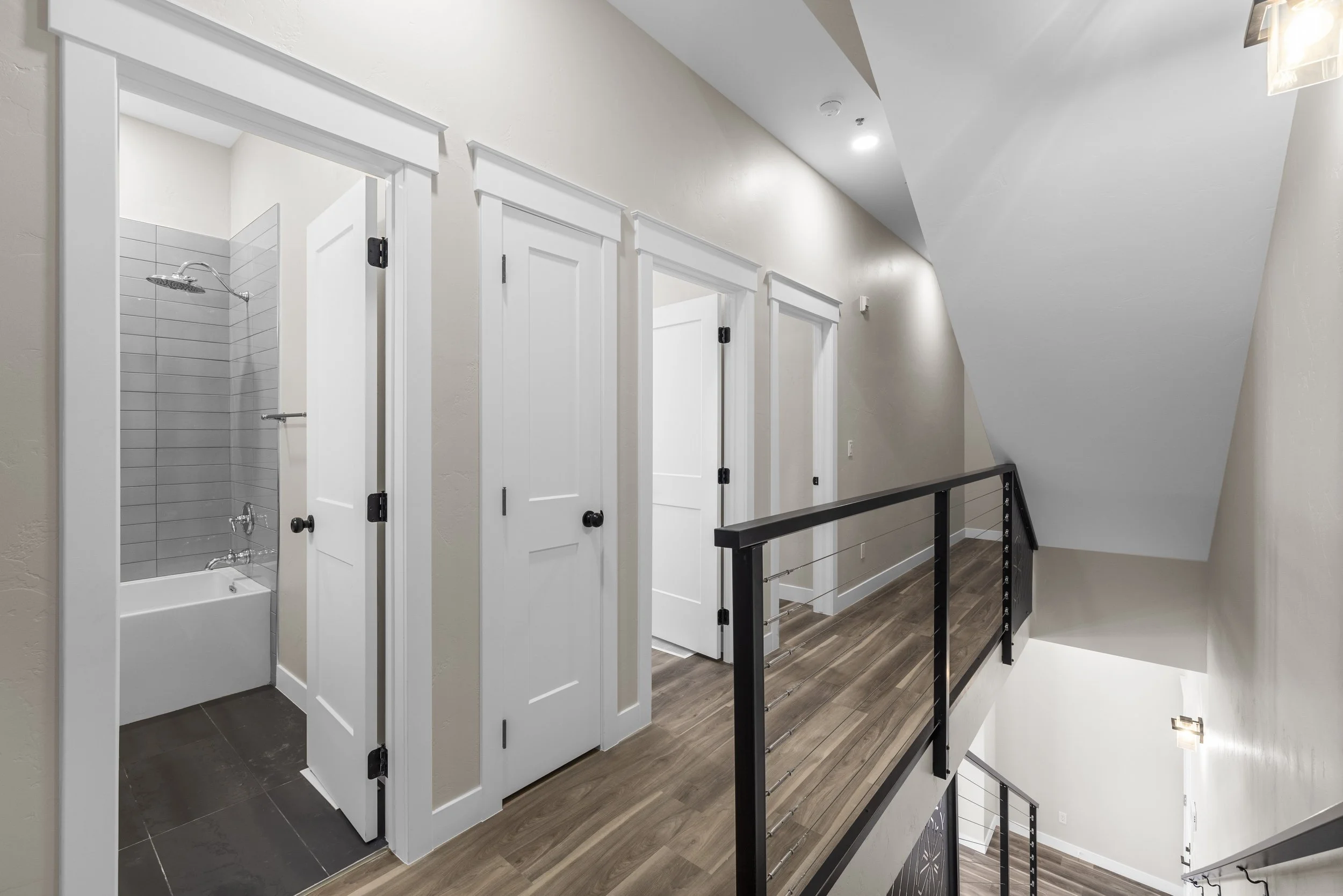 A modern home's upper hallway with four closed white doors, a bathroom with a bathtub and shower visible on the left, wood flooring, a black metal railing, and a staircase descending to a lower level.