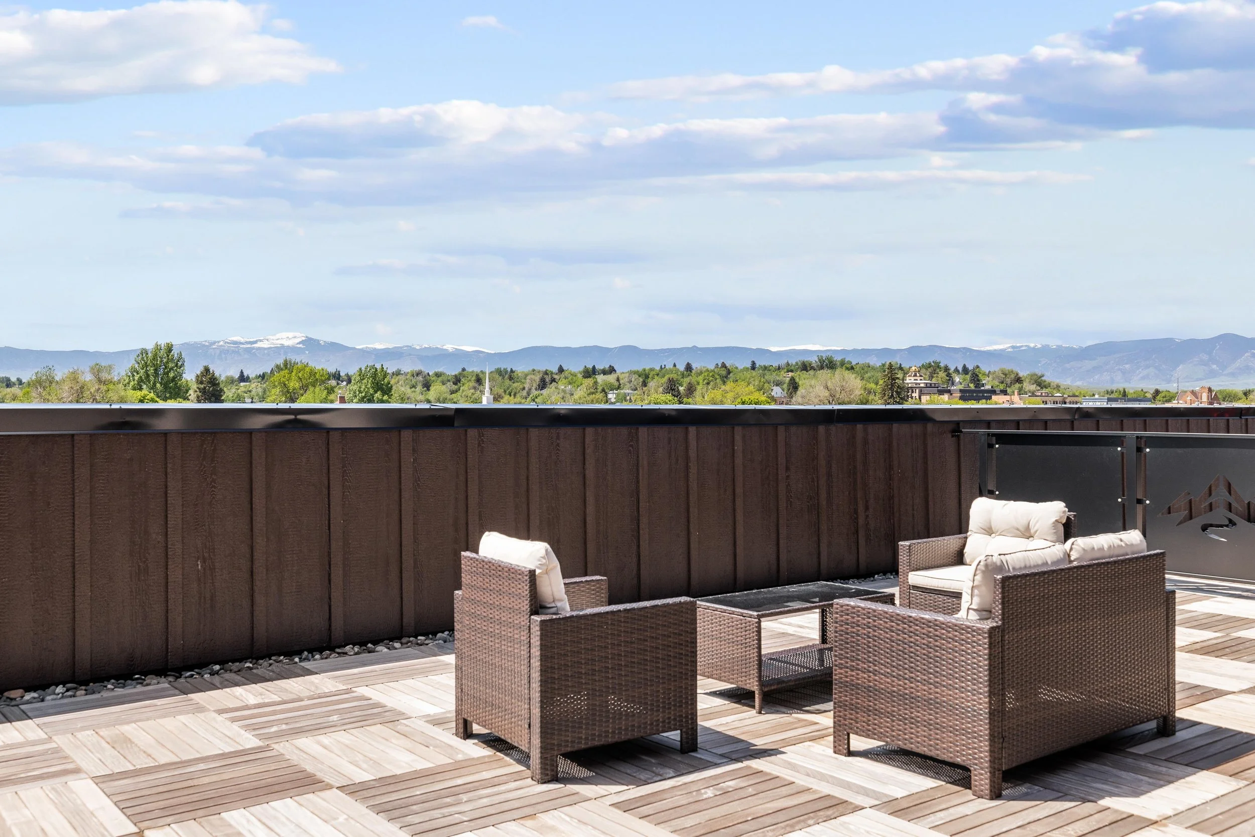 Empty outdoor rooftop patio in an Ice Haus Condo for sale in Sheridan, Wyoming, with brown wicker furniture, beige cushions, and a view of green trees, mountains, and a cloudy sky.