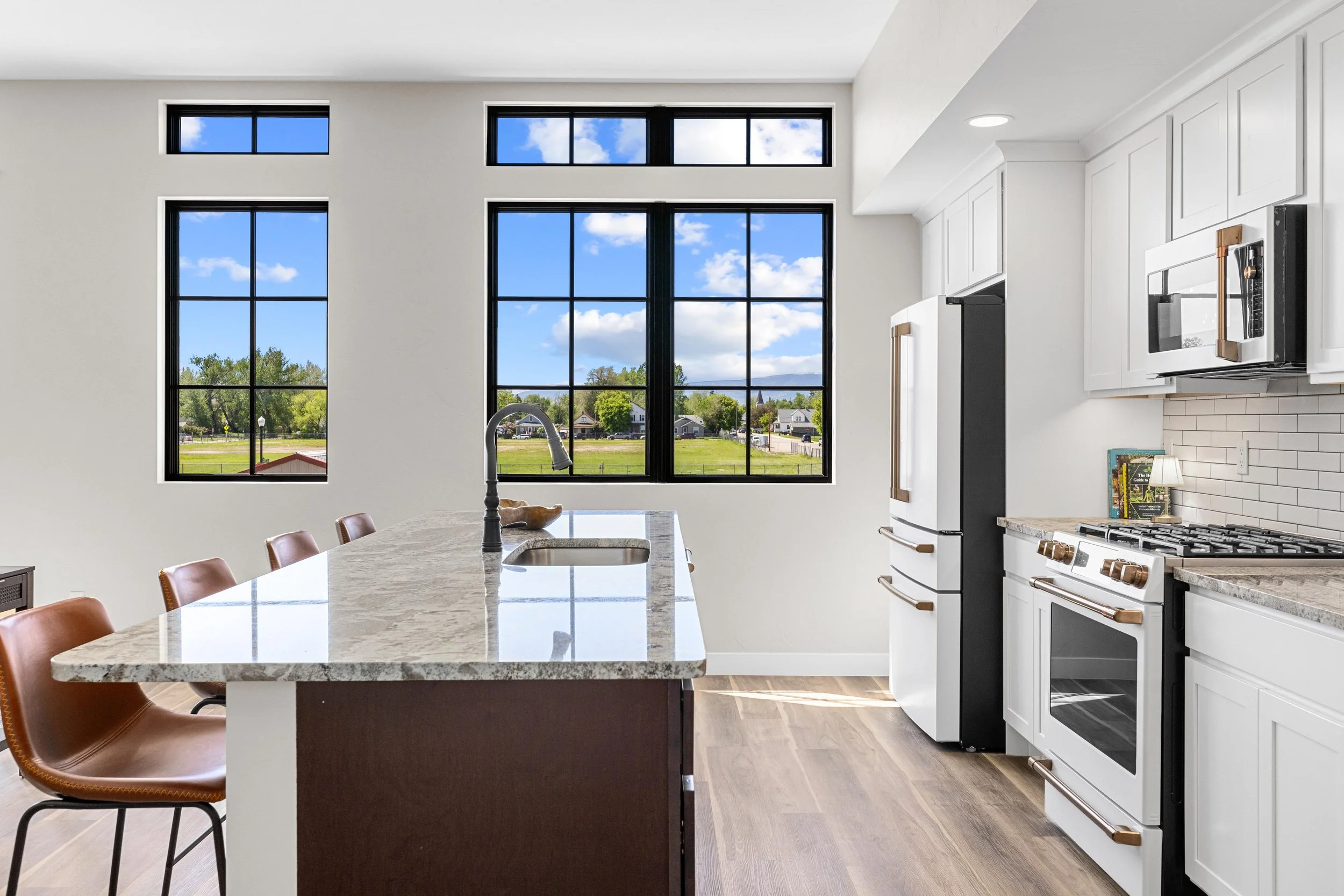 Modern kitchen with large black-framed windows, white cabinets, granite countertops, and stainless steel appliances, overlooking a green outdoor landscape.
