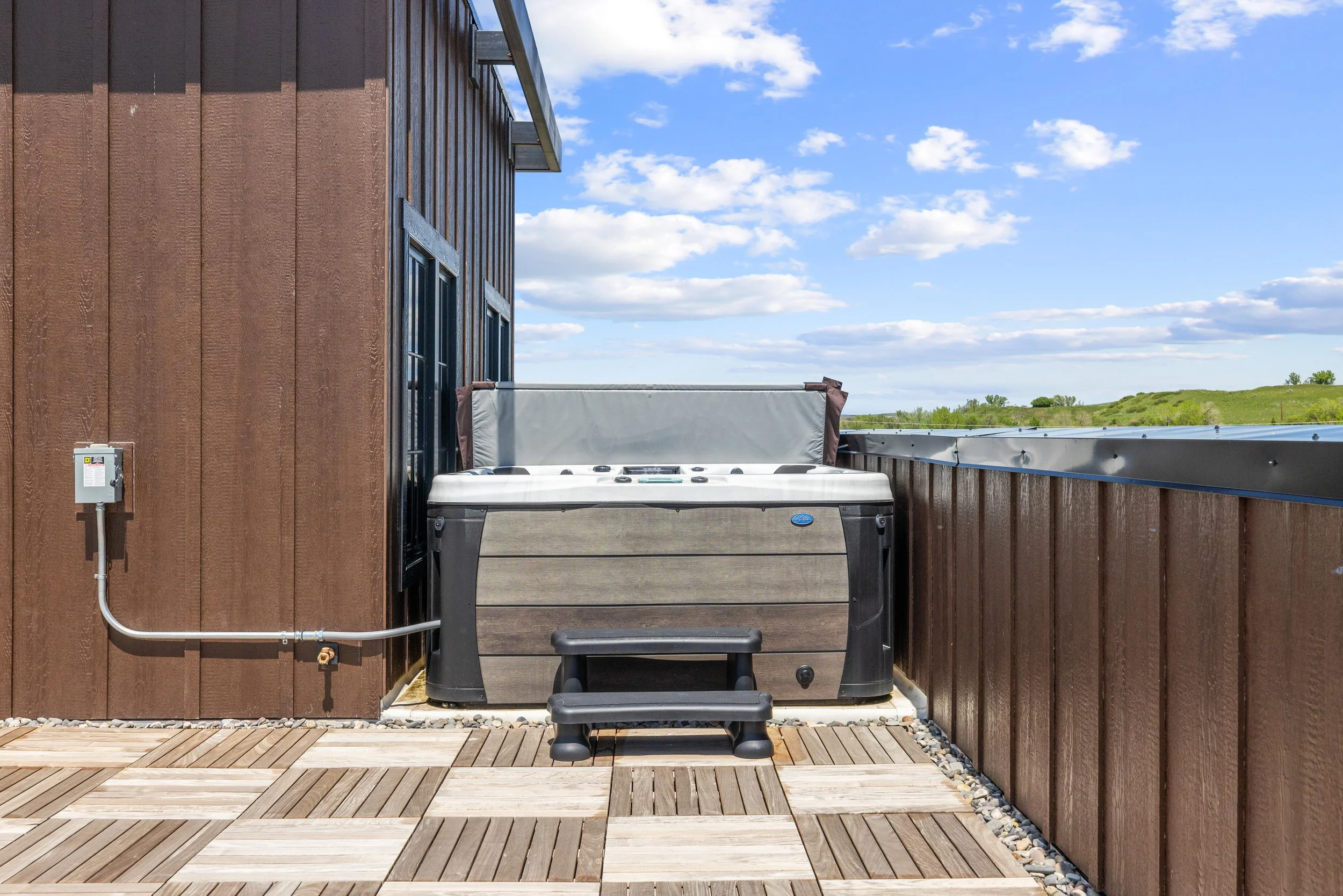 Outdoor hot tub on a wooden deck next to a building with a brown exterior wall, overlooking grassy fields under a partly cloudy sky.