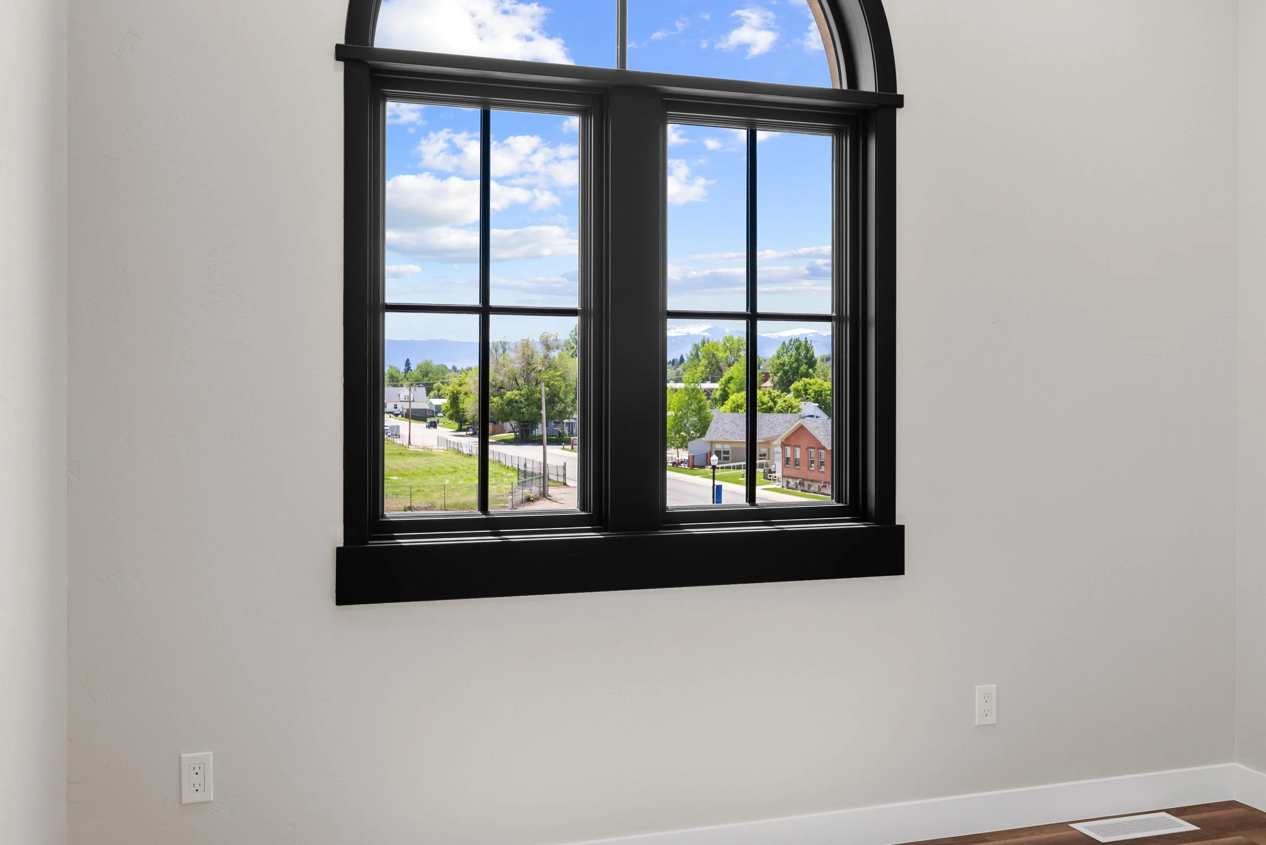 View through a large, black-framed window showing a residential neighborhood with green trees, houses, and mountains under a partly cloudy sky.